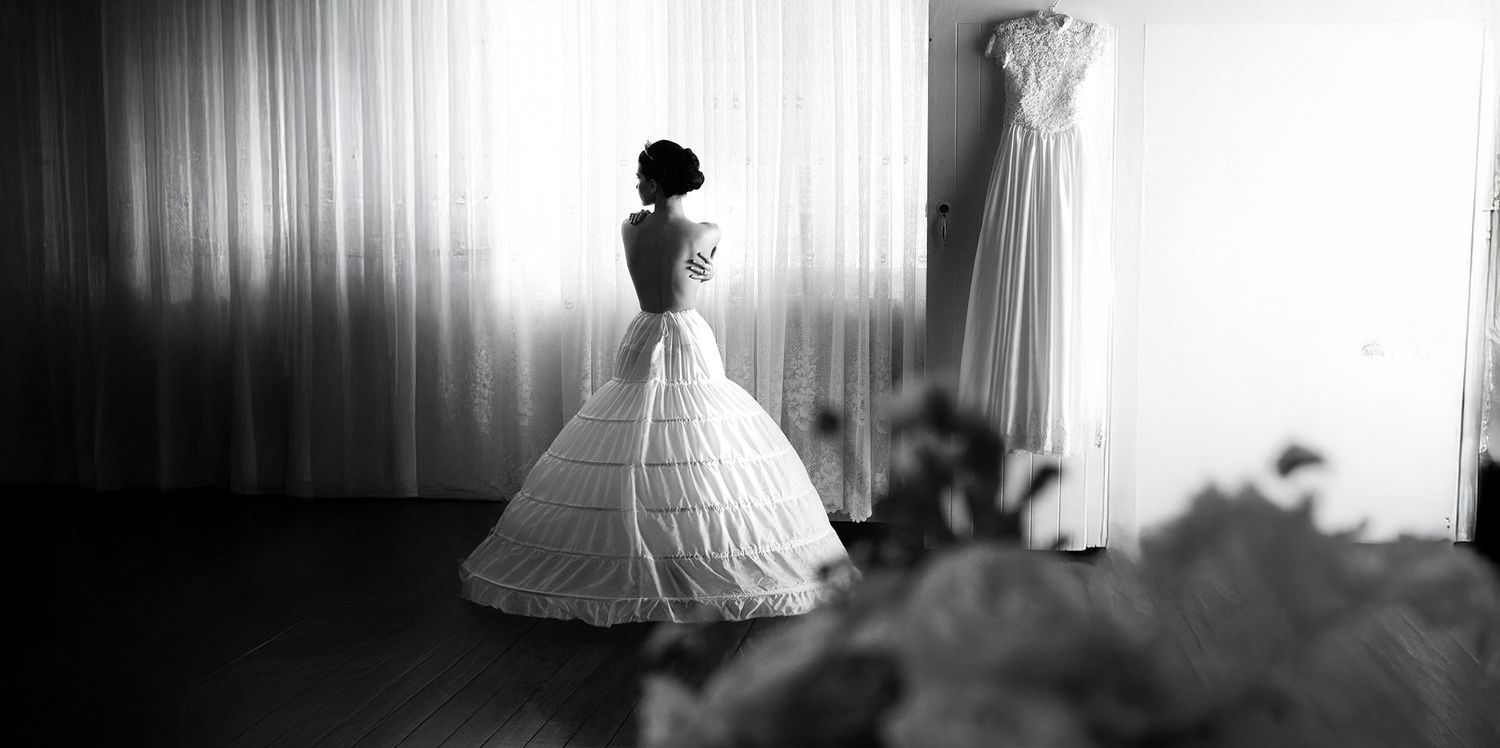 Black and white photo of a bride in a full ball gown standing before sheer curtains in a bright room.