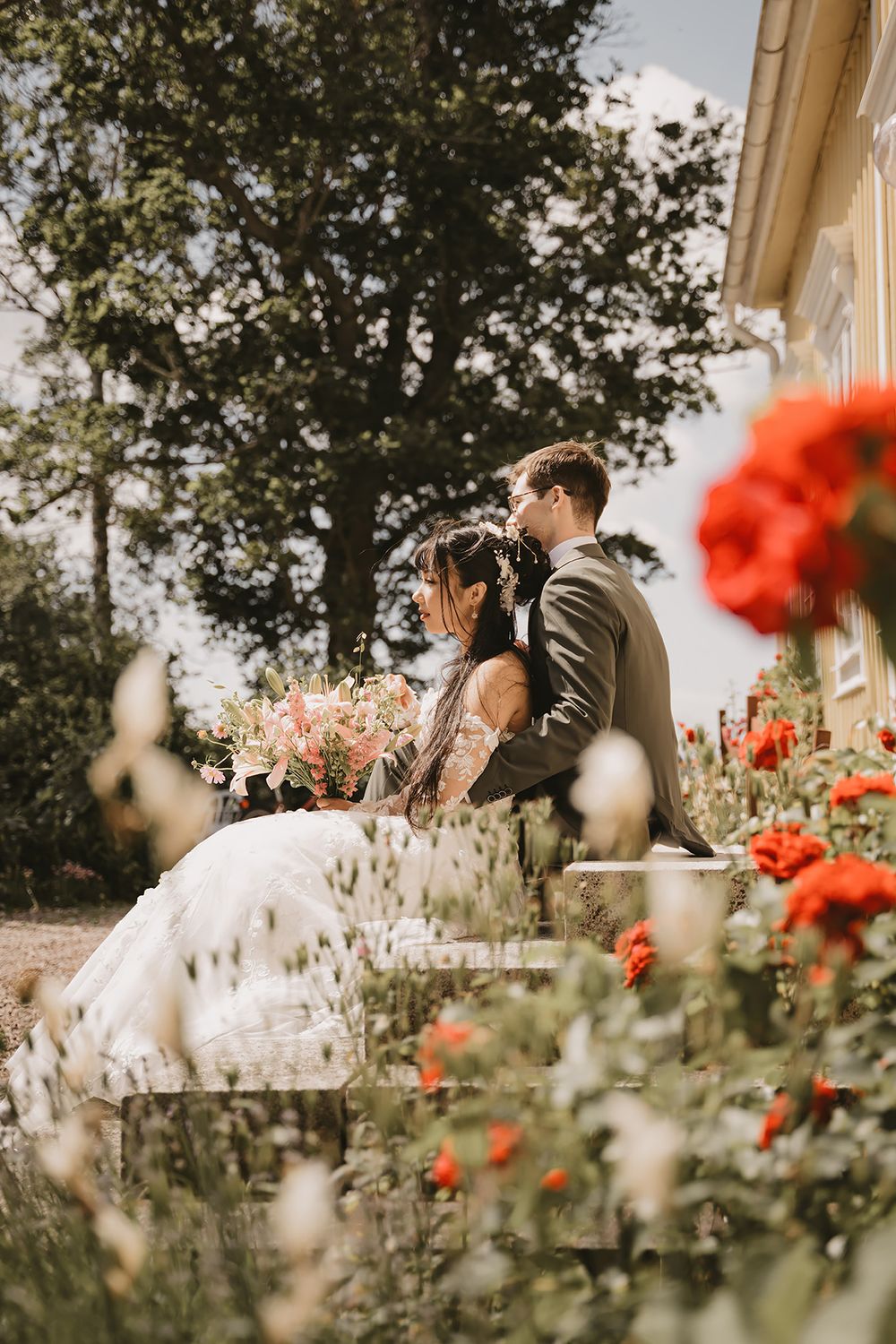 Bröllopspar sitter på en trappa, i förgrunden syns vackra röda rosor. Bröllopsfoto av Lets Bloom Media i Östergötland.