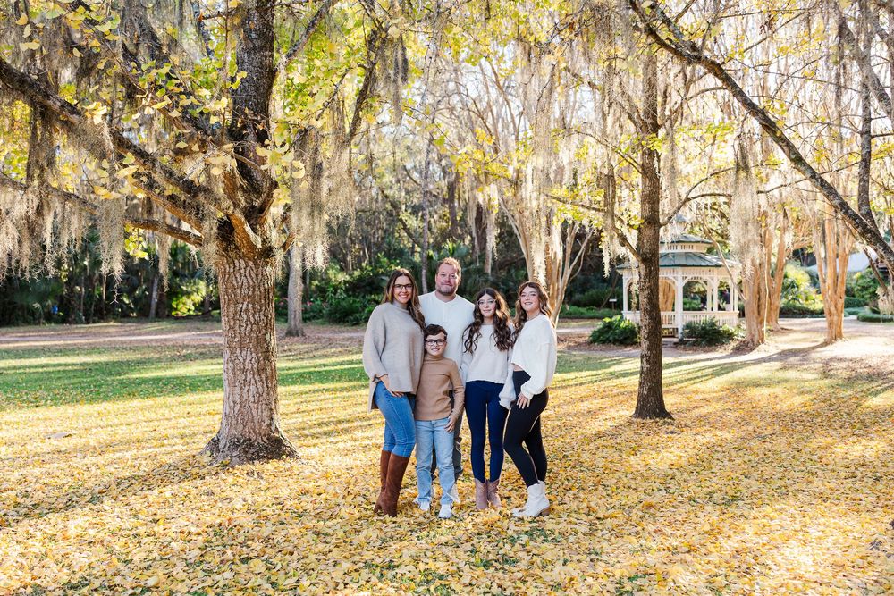 Family members pose together on fallen autumn leaves in a park setting with golden sunlight filtering through trees.