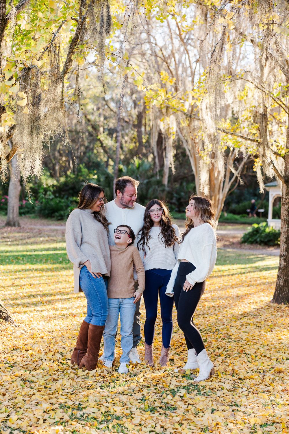 Group laughs together while standing on golden fall leaves under moss-covered trees in a park.