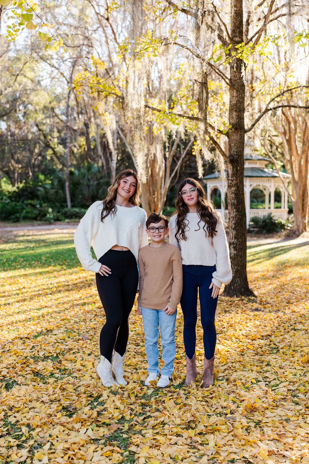 Three people laugh together while standing on fallen autumn leaves in a sunlit park.