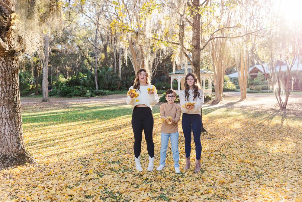 Three friends stand together on fallen autumn leaves under moss-draped trees in a sunlit park setting.