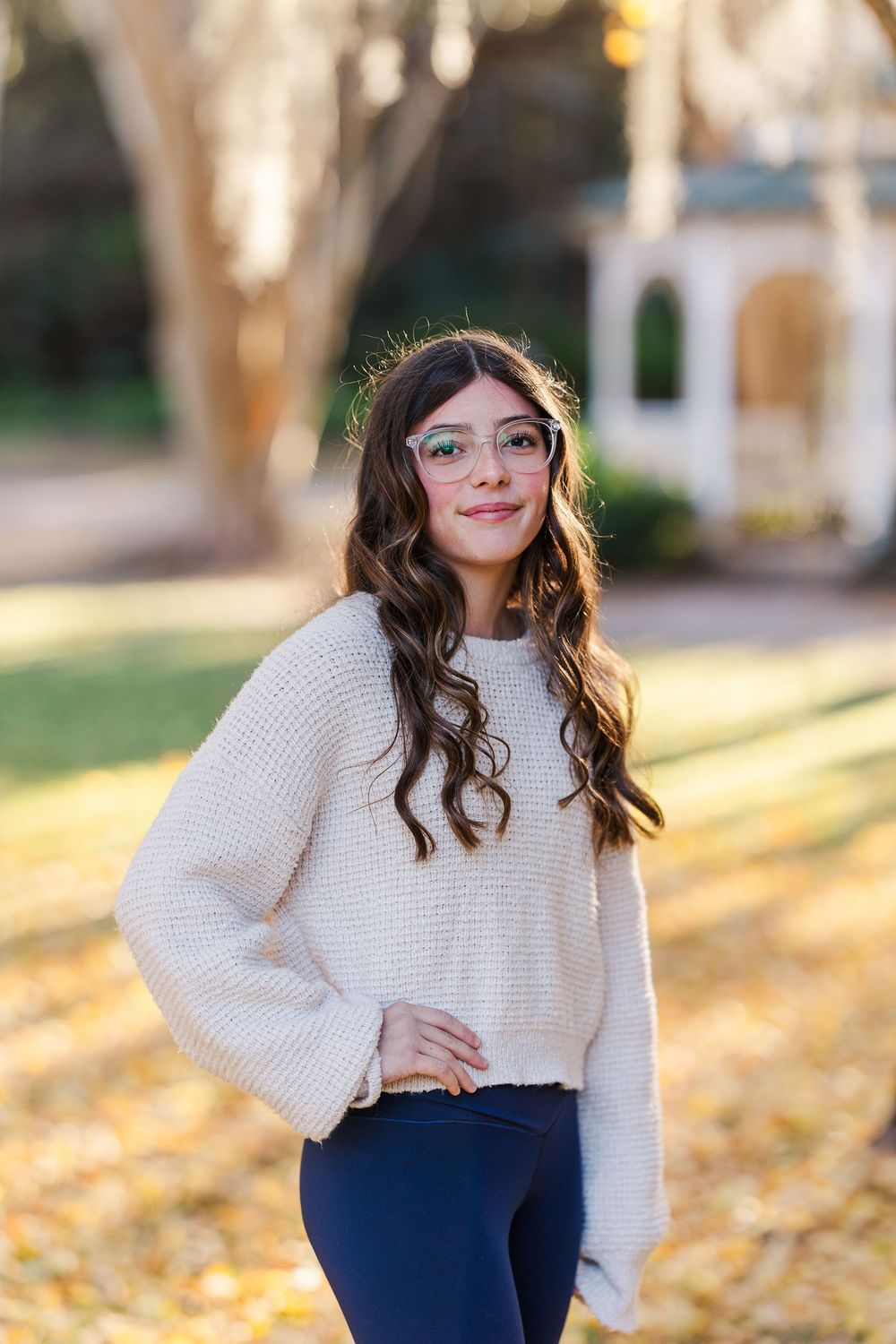 Woman in cream sweater and dark pants poses outdoors during autumn with golden sunlight filtering through trees.