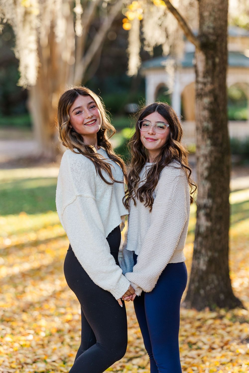 Two friends in cozy sweaters share joyful moments among fallen autumn leaves and trees.