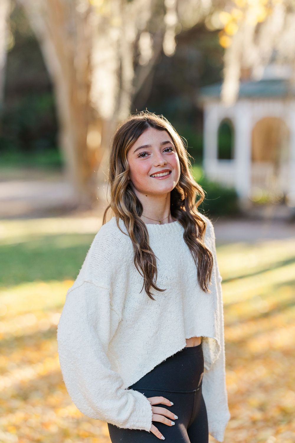 A person wearing a white sweater and black top poses outdoors in autumn with trees in the background.