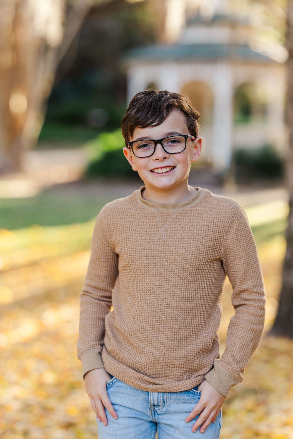 A child in a tan sweater smiles while standing in autumn leaves.