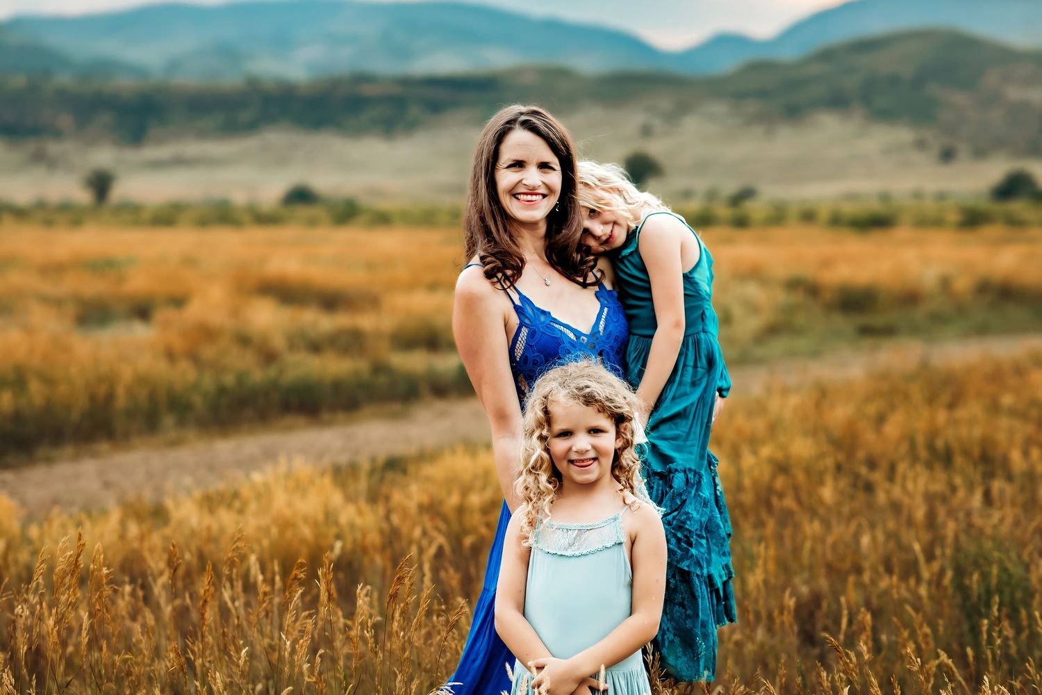 Three people in blue dresses stand together in a golden field with mountains in the background.