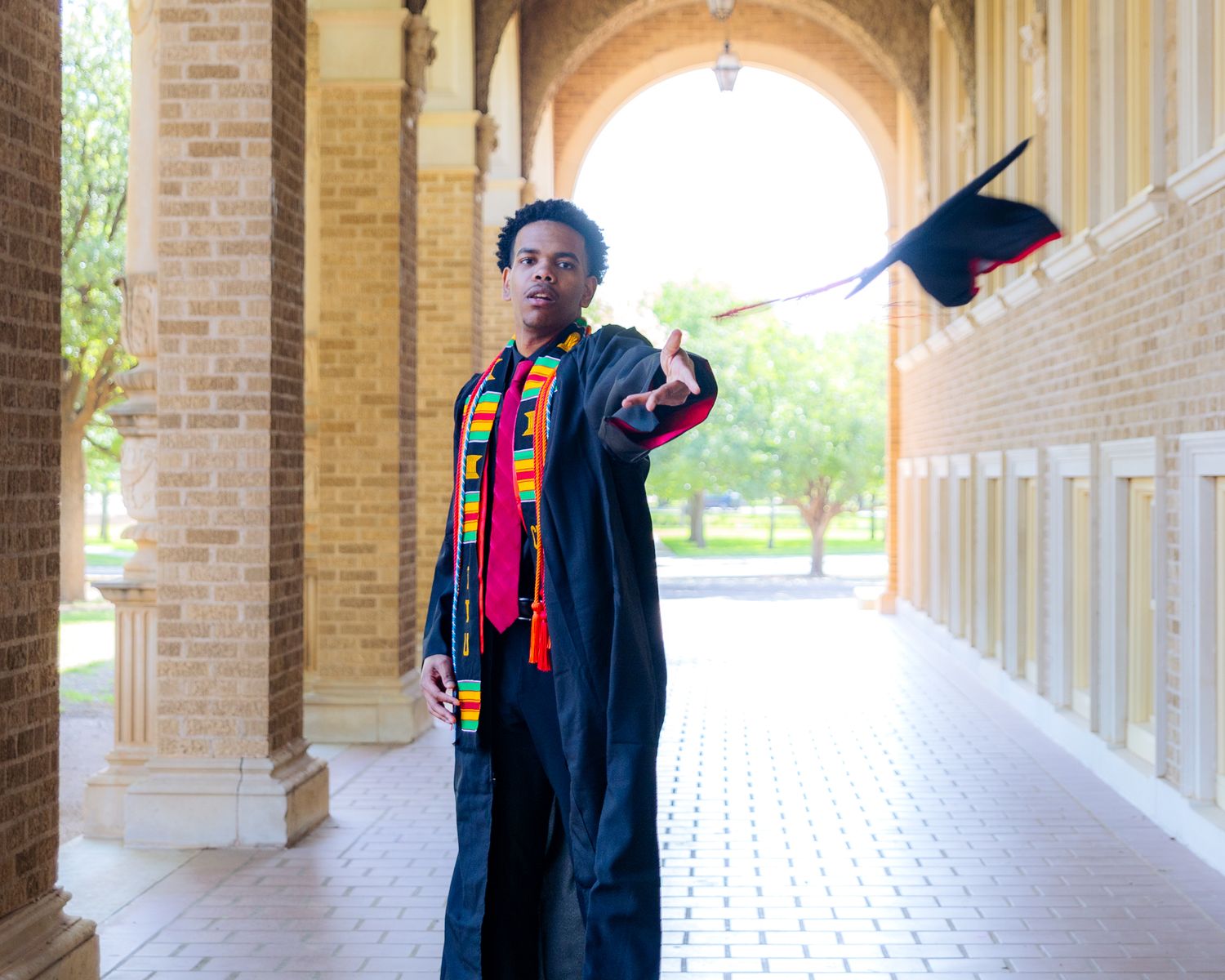 Graduate celebrating by tossing cap in a bright outdoor hallway, wearing a colorful stole and gown capturing achievement, joy, and graduate