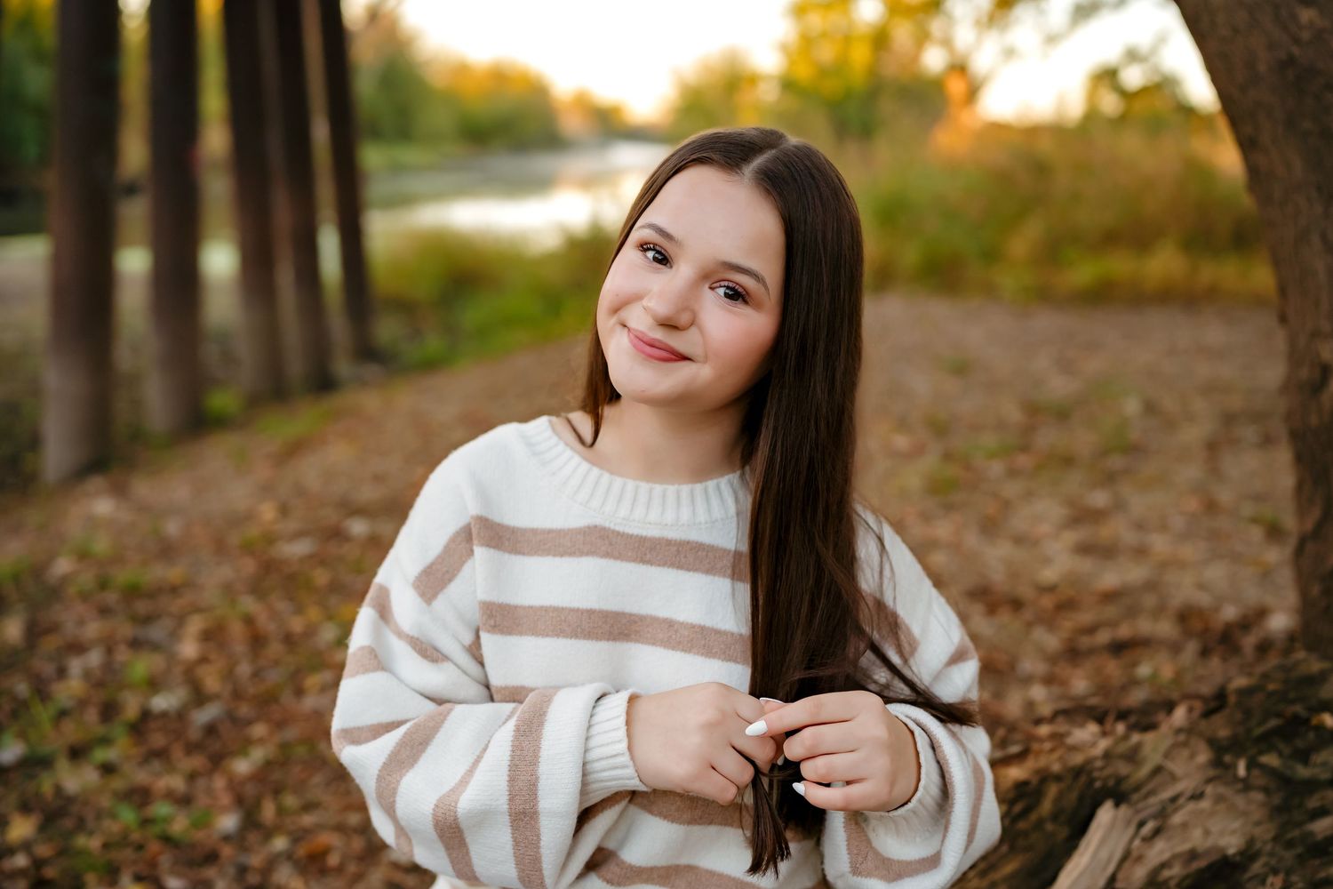 A person in a striped sweater poses outdoors during autumn with golden foliage and trees in the background.