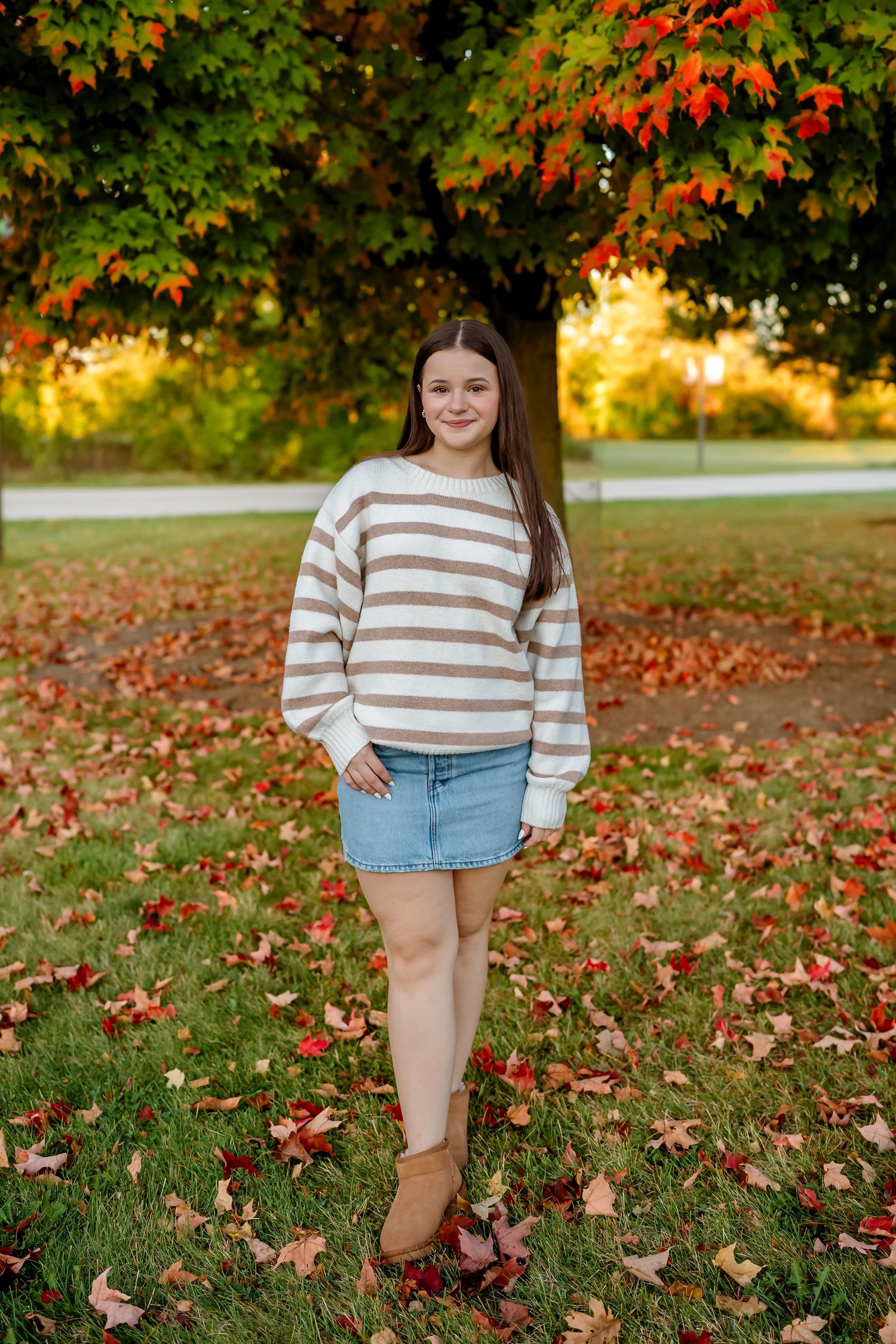 Fall portrait session among colorful maple leaves on grass wearing a cozy striped sweater and jean shorts.