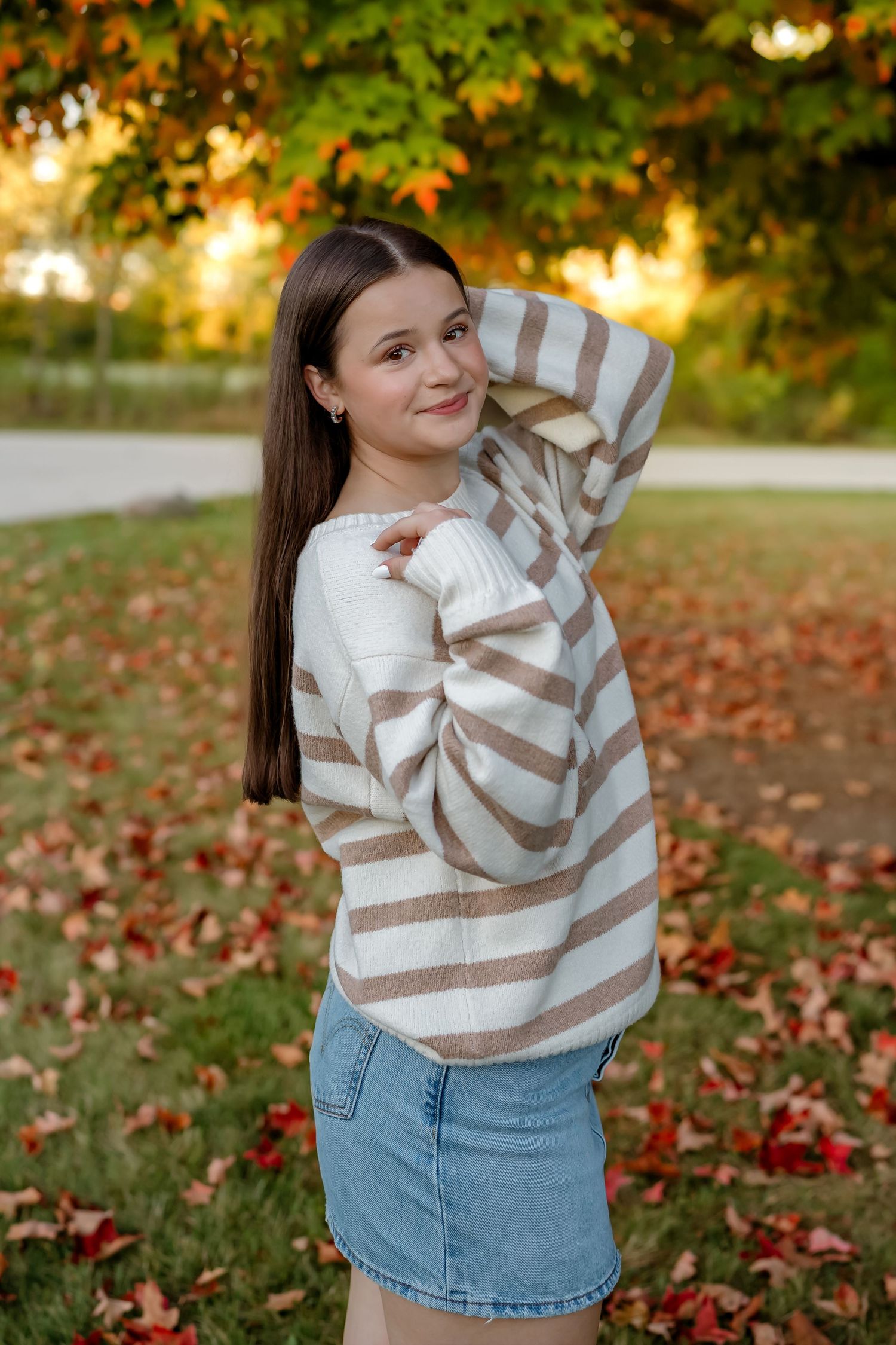A woman in a striped sweater and jeans poses playfully in a park with autumn leaves scattered on the ground.