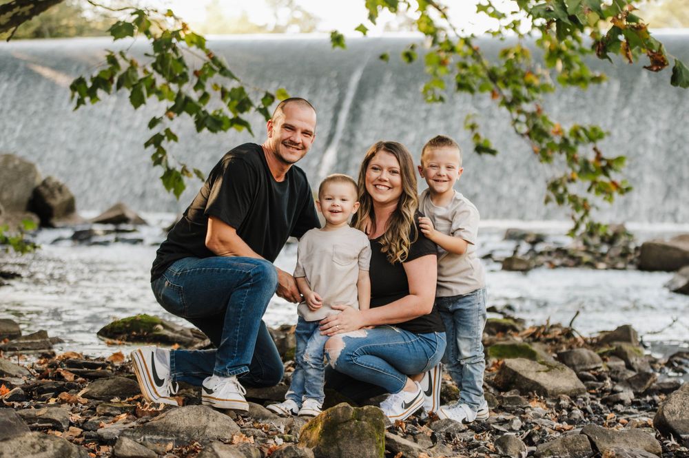 Family enjoying a casual outdoor photoshoot on rocky riverbank surrounded by green foliage.