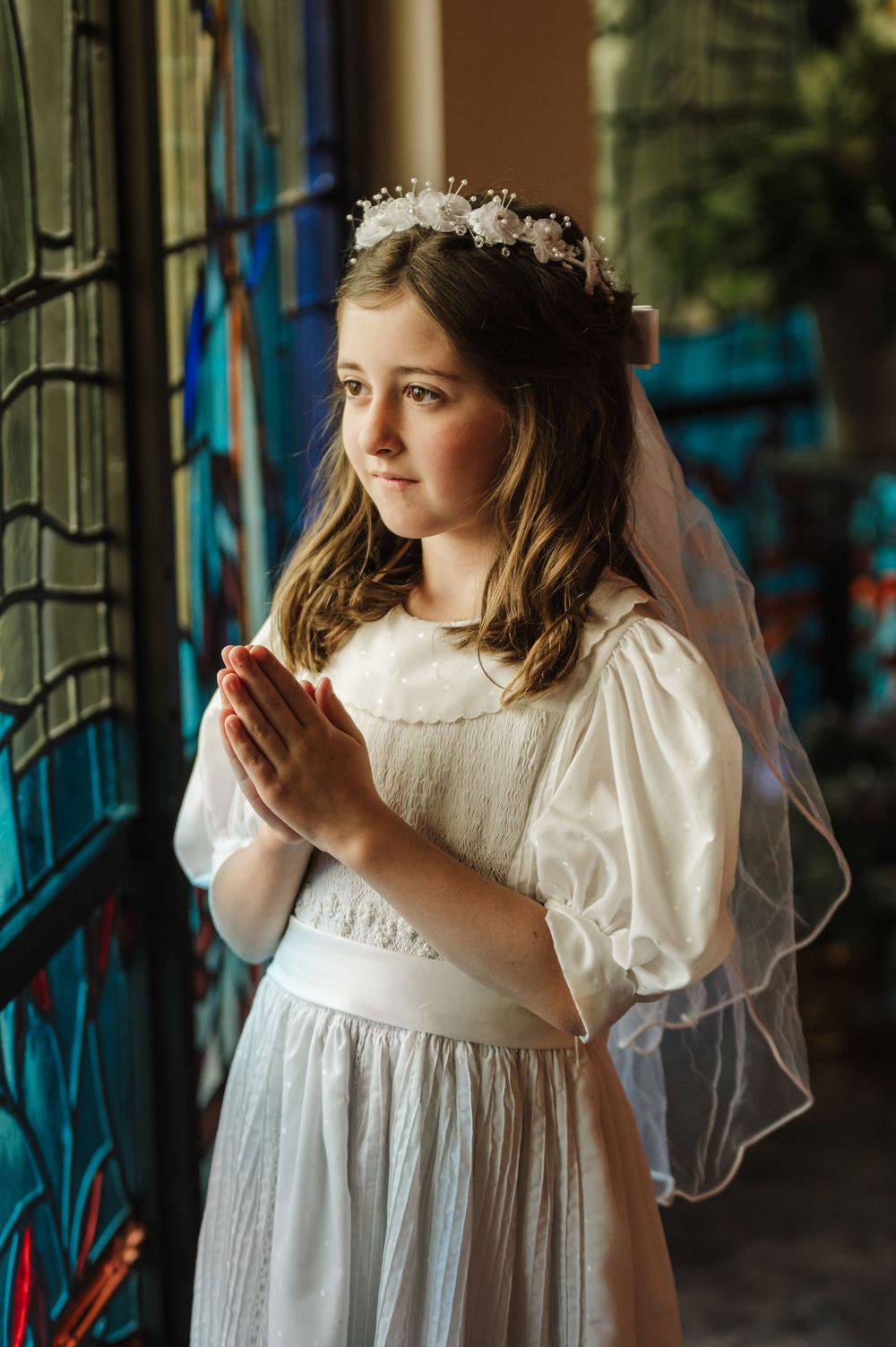 Young person in white communion dress poses near stained glass windows.