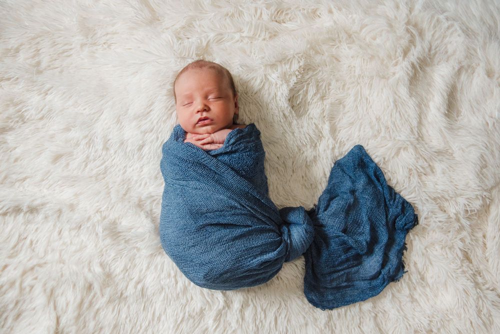 Newborn baby wrapped in blue swaddle on white textured backdrop.