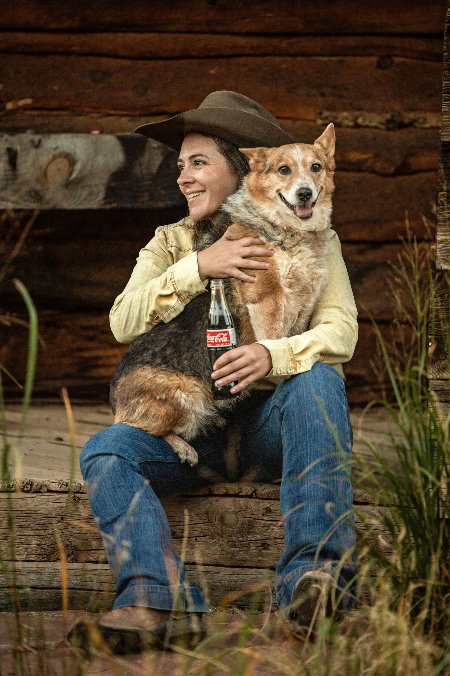 Person in western attire holding a happy corgi dog and coca-cola bottle against rustic wooden backdrop.