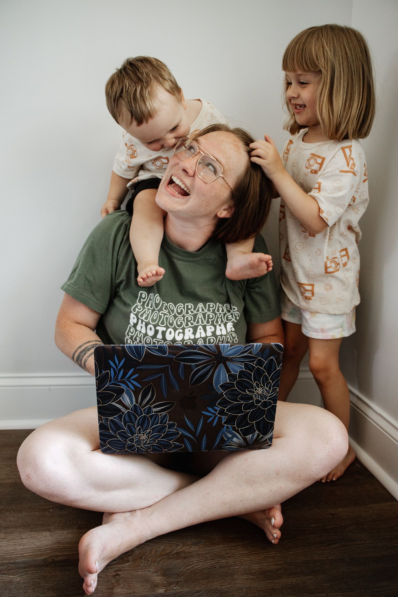A mom sitting cross-legged on floor with laptop on lap. A small boy is sitting on her shoulders, and a little girl is standing beside them.