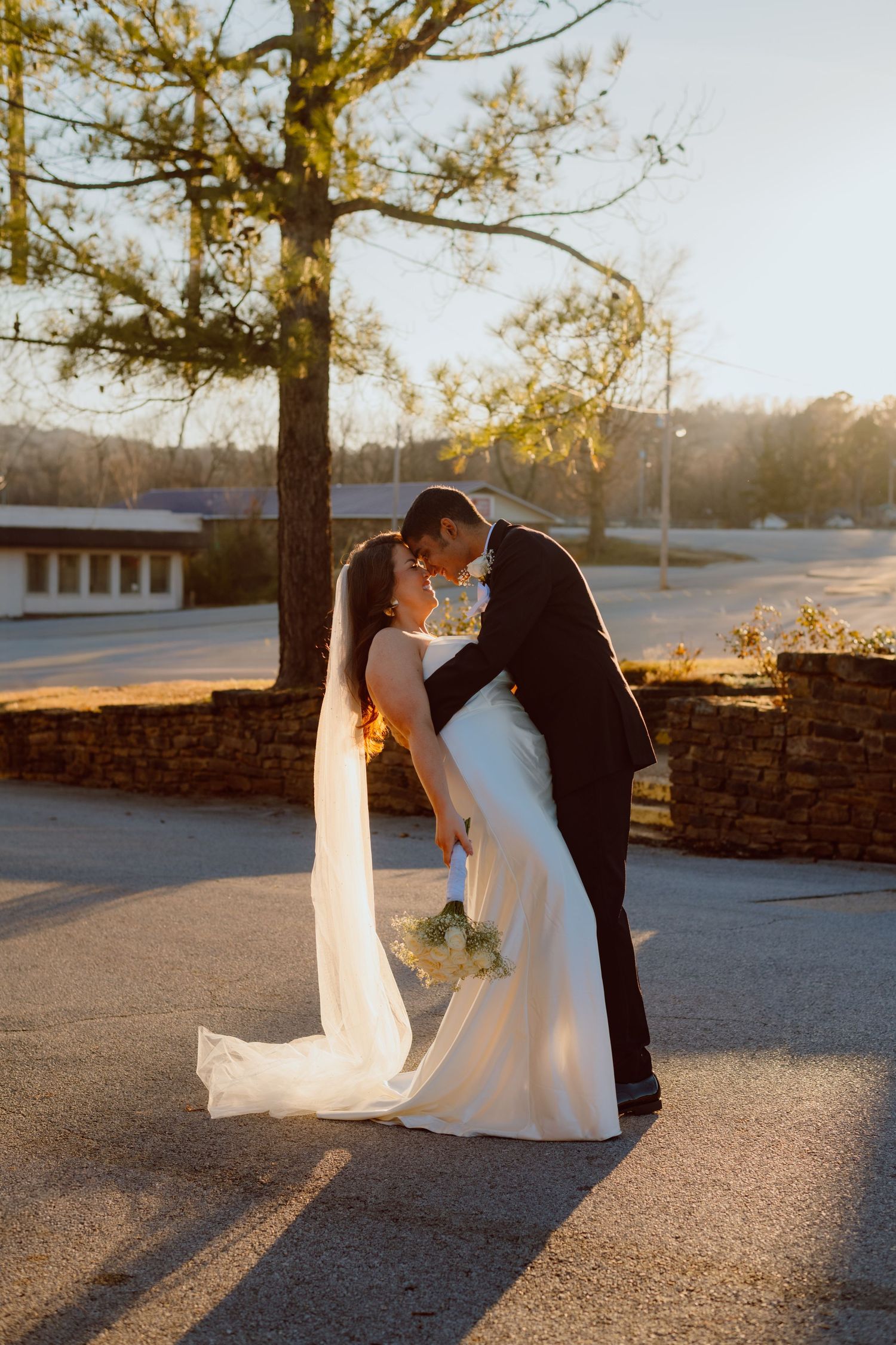 A romantic sunset wedding photo of a couple kissing while the bride holds a bouquet on a stone walkway.