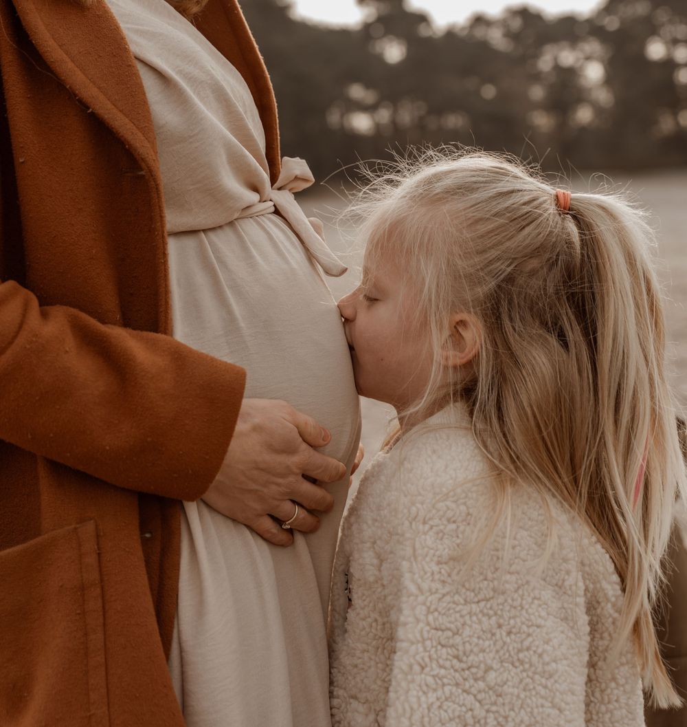 A child in a cream sweater kisses a pregnant belly while standing next to someone wearing a brown coat.