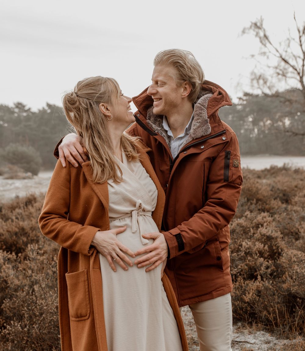 A couple in brown coats share an intimate moment outdoors during a maternity photoshoot at sunset.