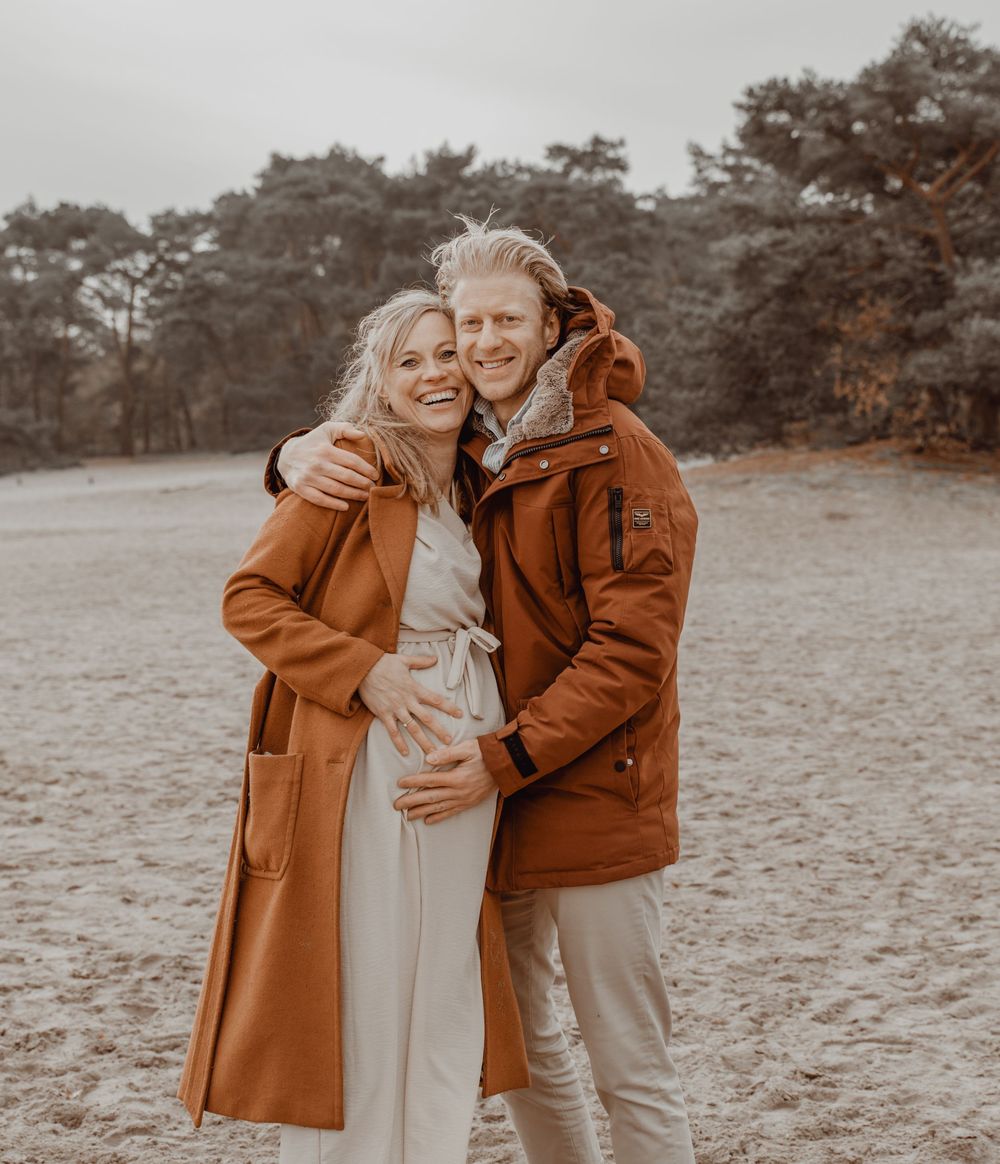 A couple wearing brown coats embraces while standing in a grassy field with pine trees in the background.