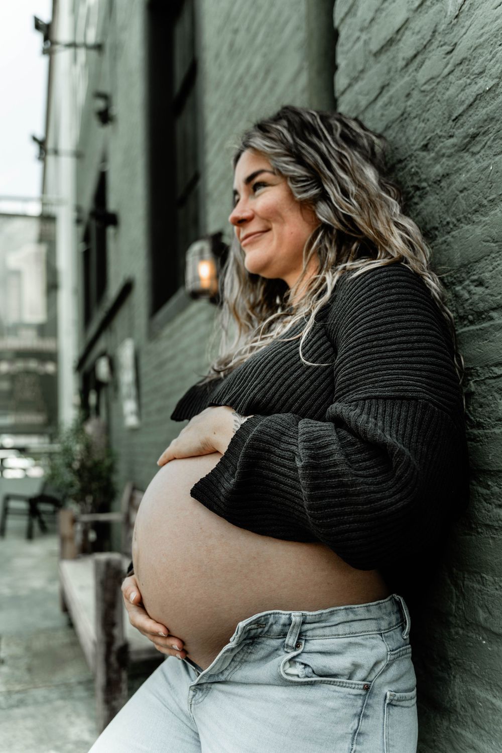 Pregnant woman in black sweater smiles while leaning against industrial green wall.