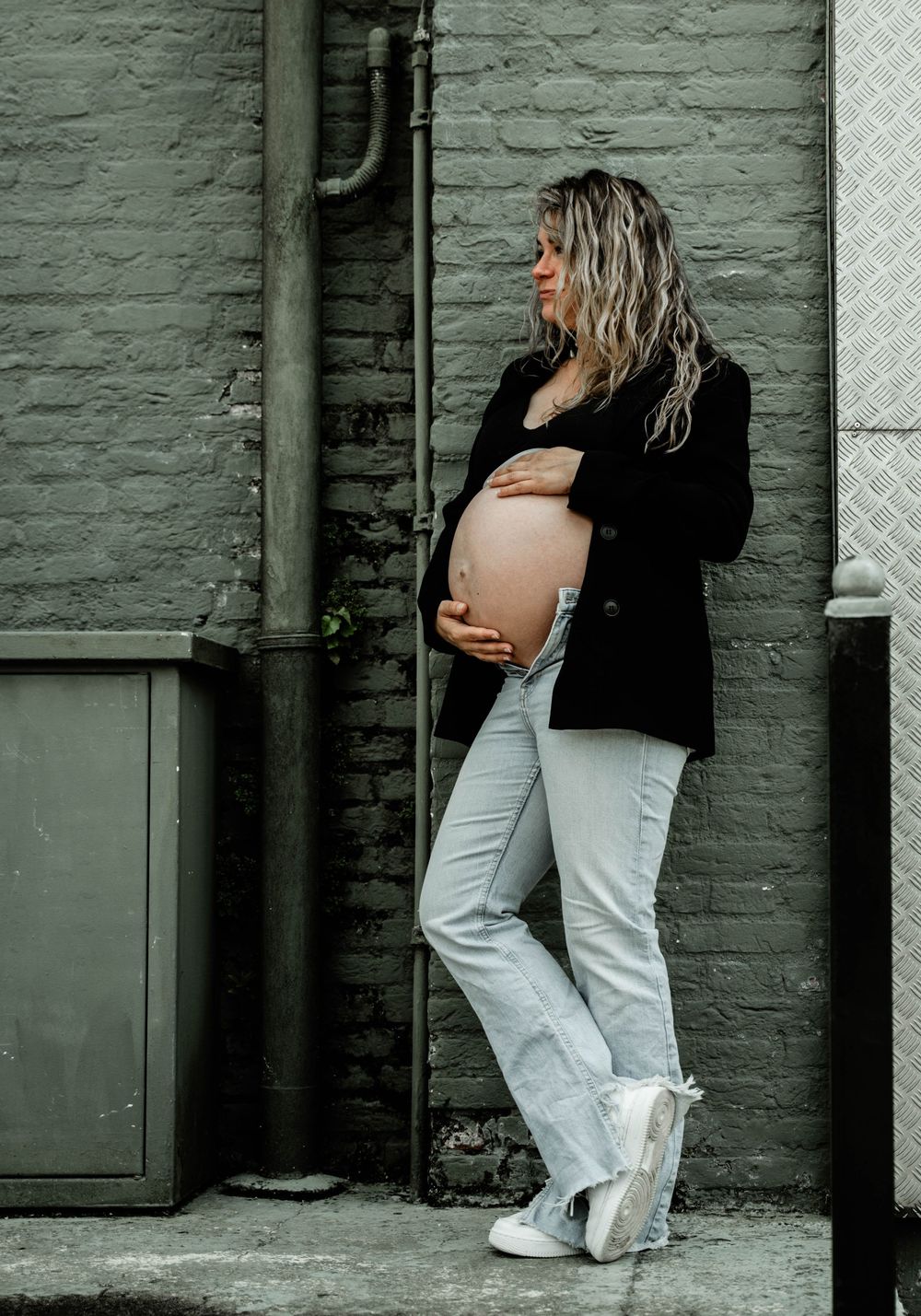 Expectant mother in black jacket and white jeans poses against green brick wall.