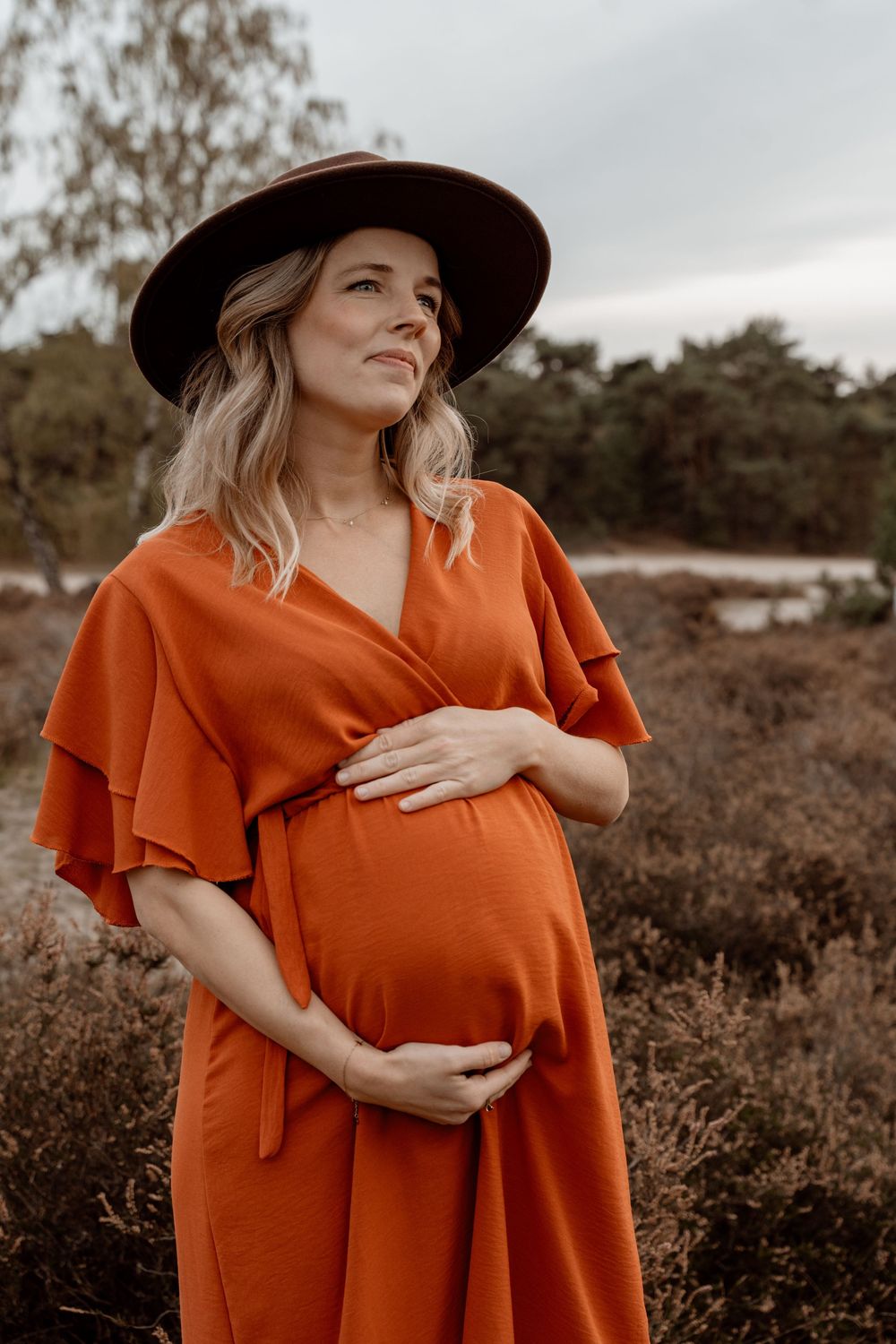 Expectant woman in a flowing rust-colored dress poses outdoors in a natural setting during golden hour.