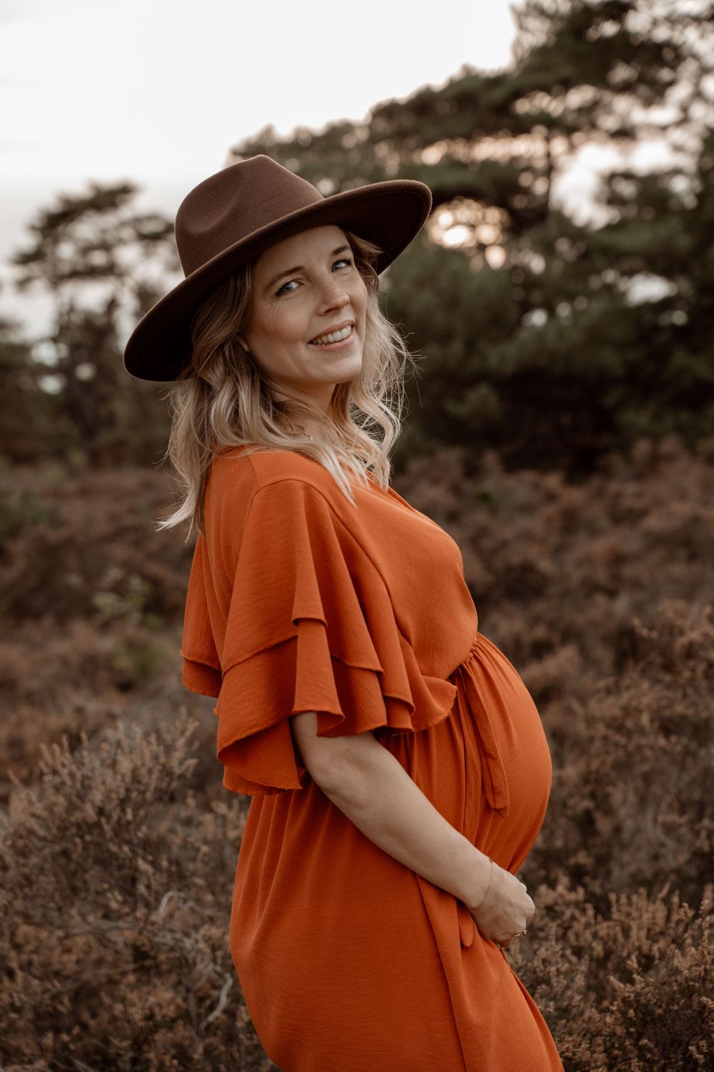 A person in an orange ruffled dress and baseball cap poses outdoors against a natural backdrop during golden hour.