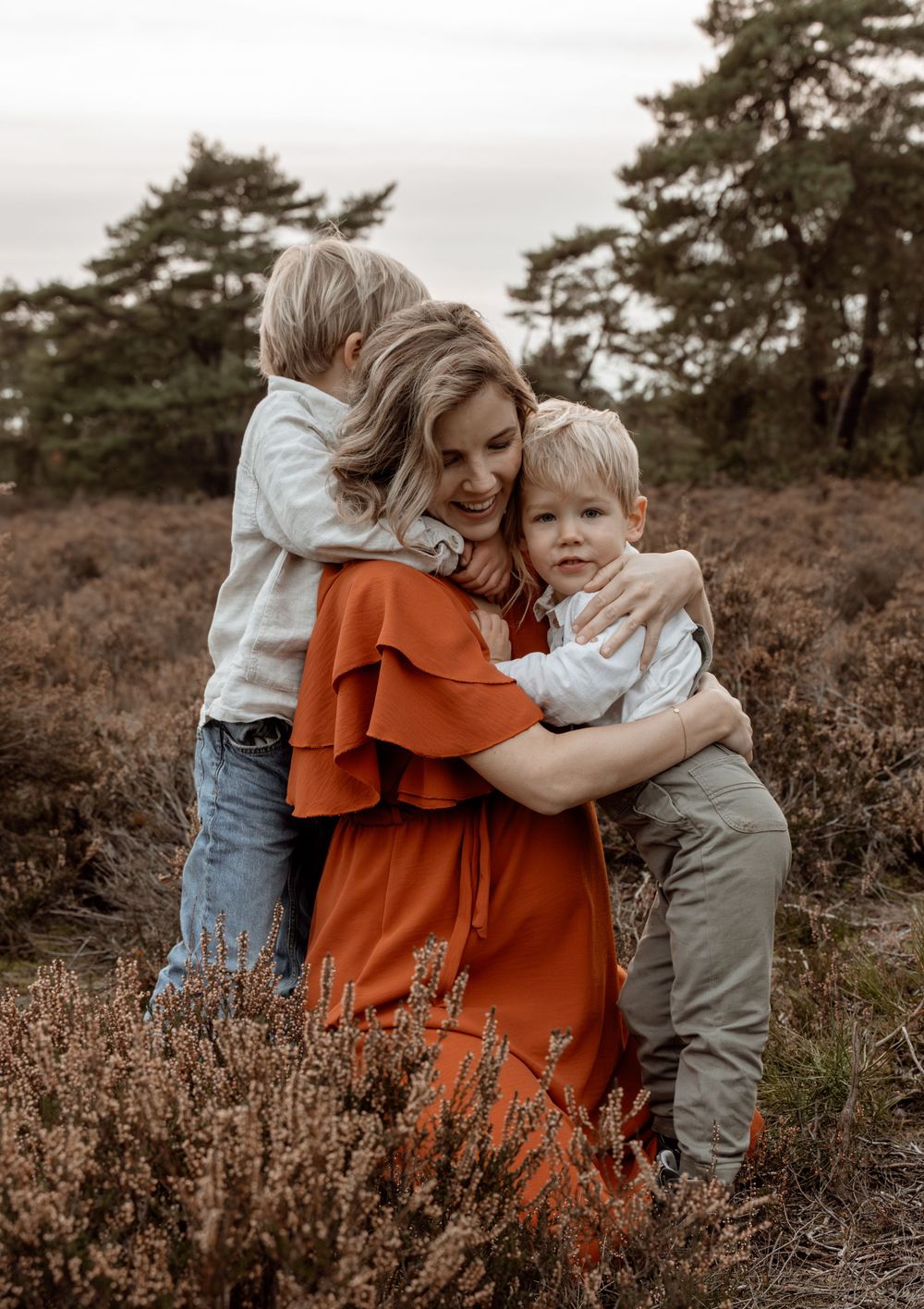 A tender embrace between figures in an orange dress and light clothing amidst a field of purple heather and pine trees.
