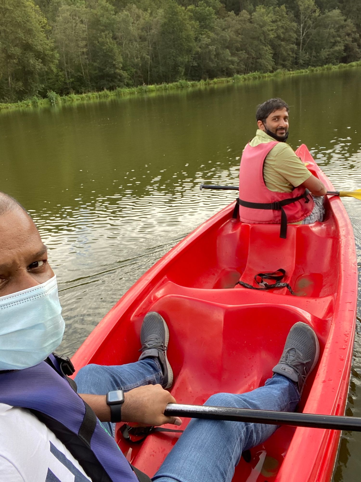 Two people wearing safety vests paddle together in a bright red kayak on a calm lake surrounded by trees.