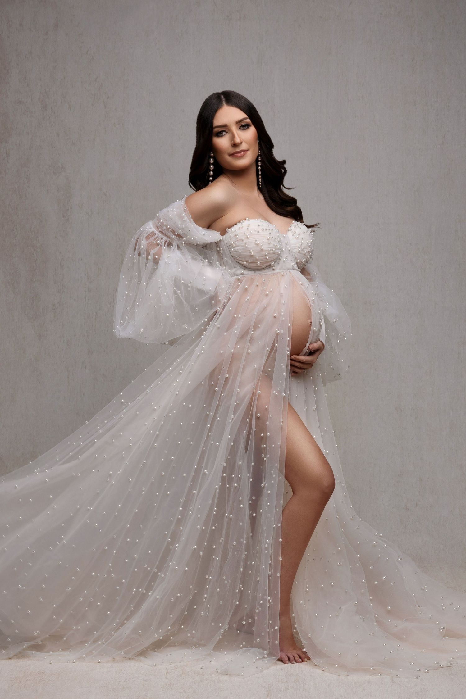Elegant maternity portrait in flowing white tulle gown with pearls against grey backdrop, showcasing flowing dark hair.