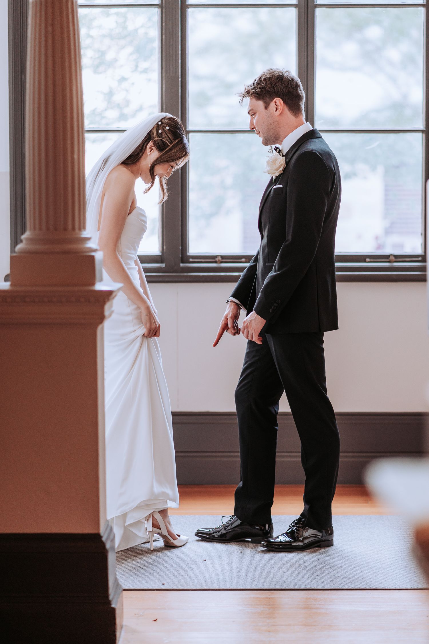 A couple at their wedding stand in front of large windows, looking at each other in formal attire.