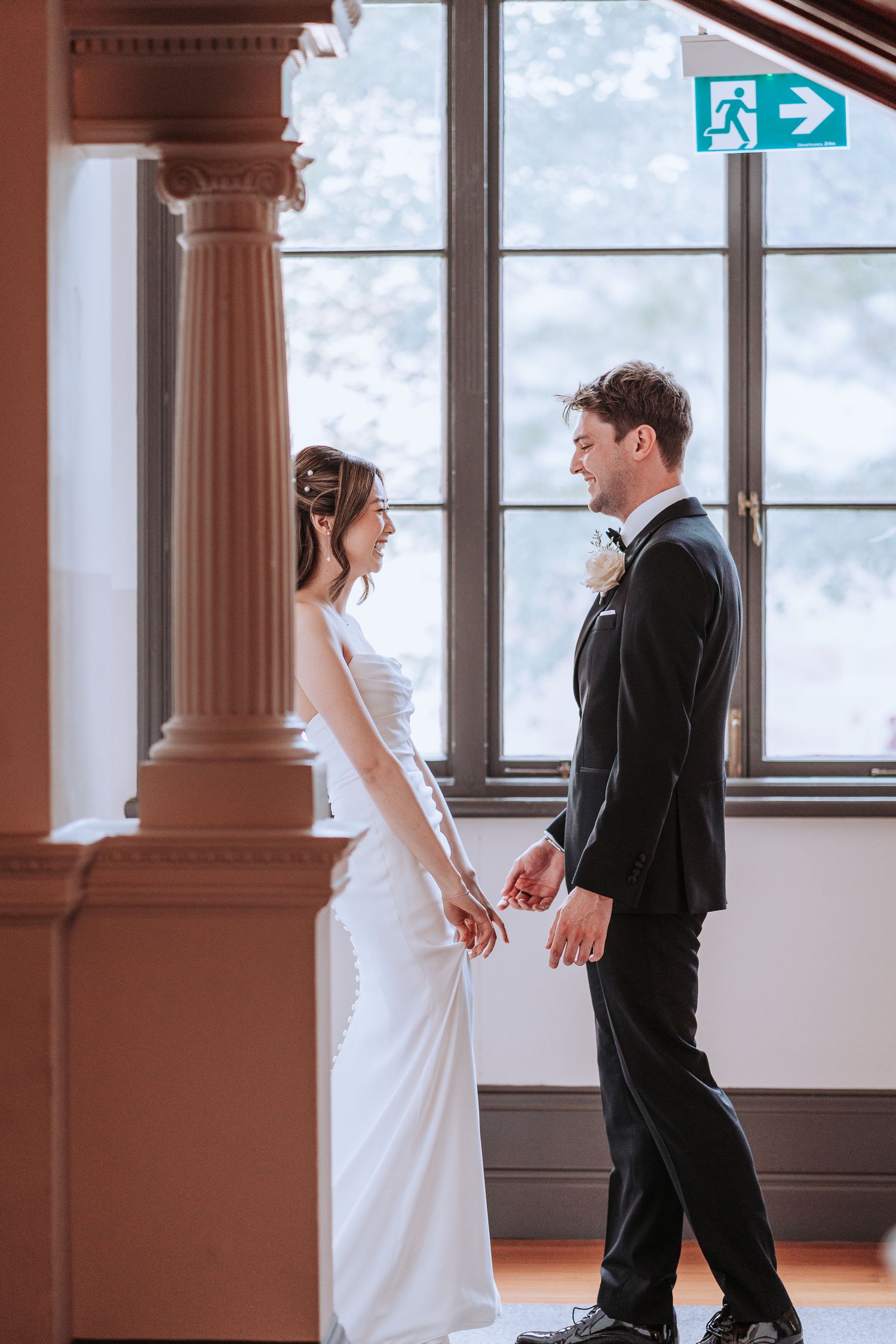 A wedding couple shares an intimate moment by a column near tall windows in formal attire.