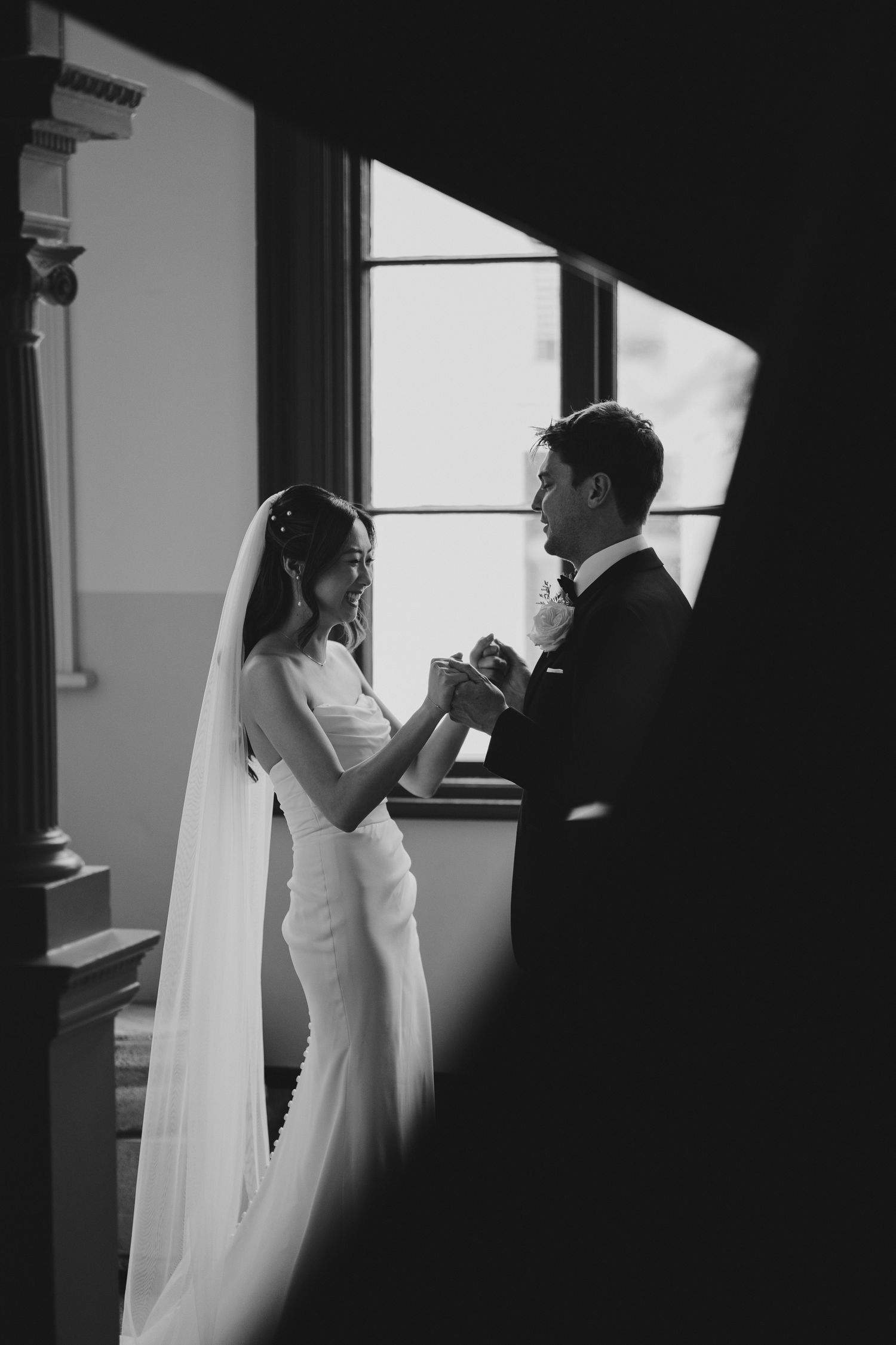 A dramatic black and white photo captures a wedding couple holding hands by a window.