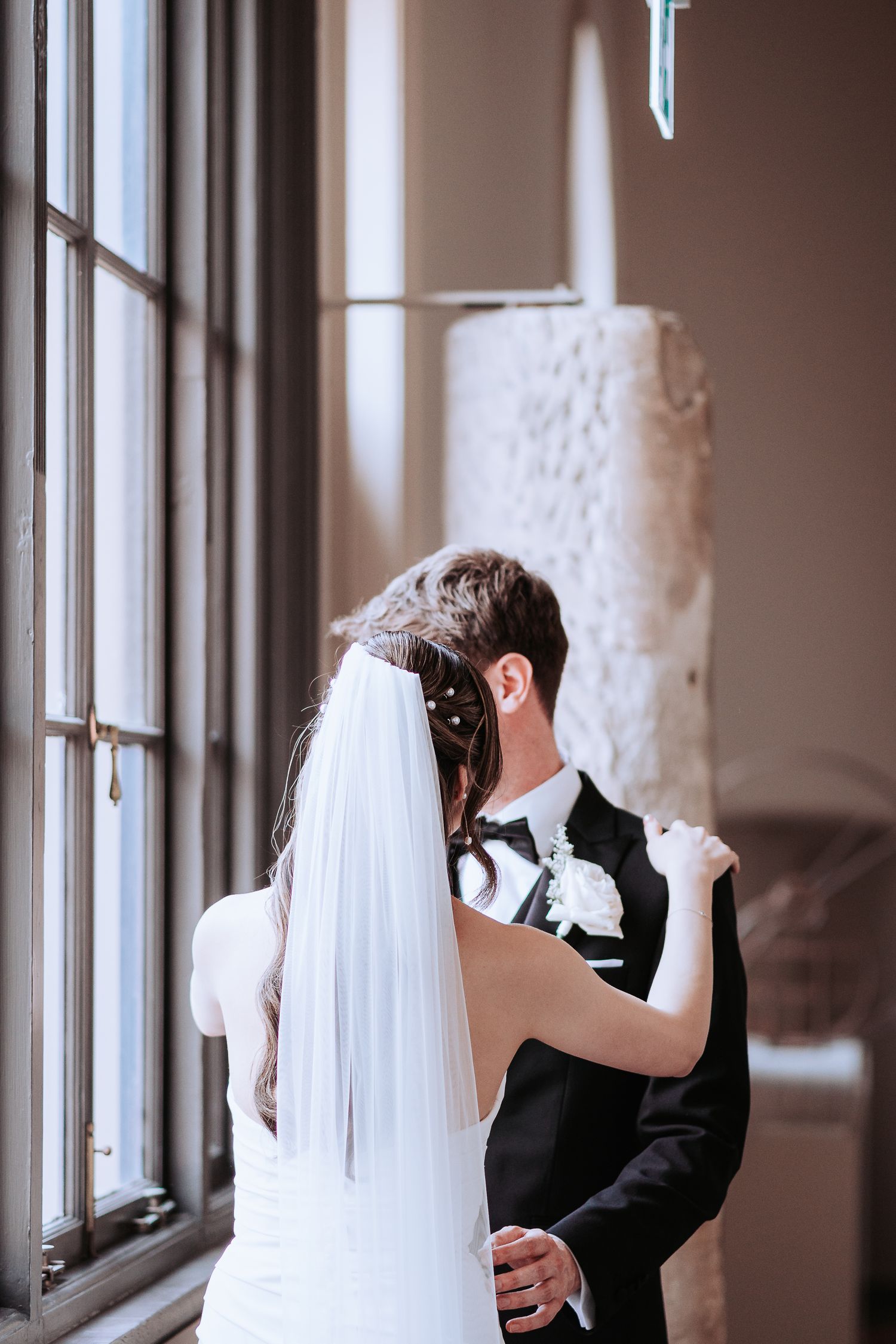 A romantic wedding moment as couple stands by window, with bride in white veil and groom in classic black suit.