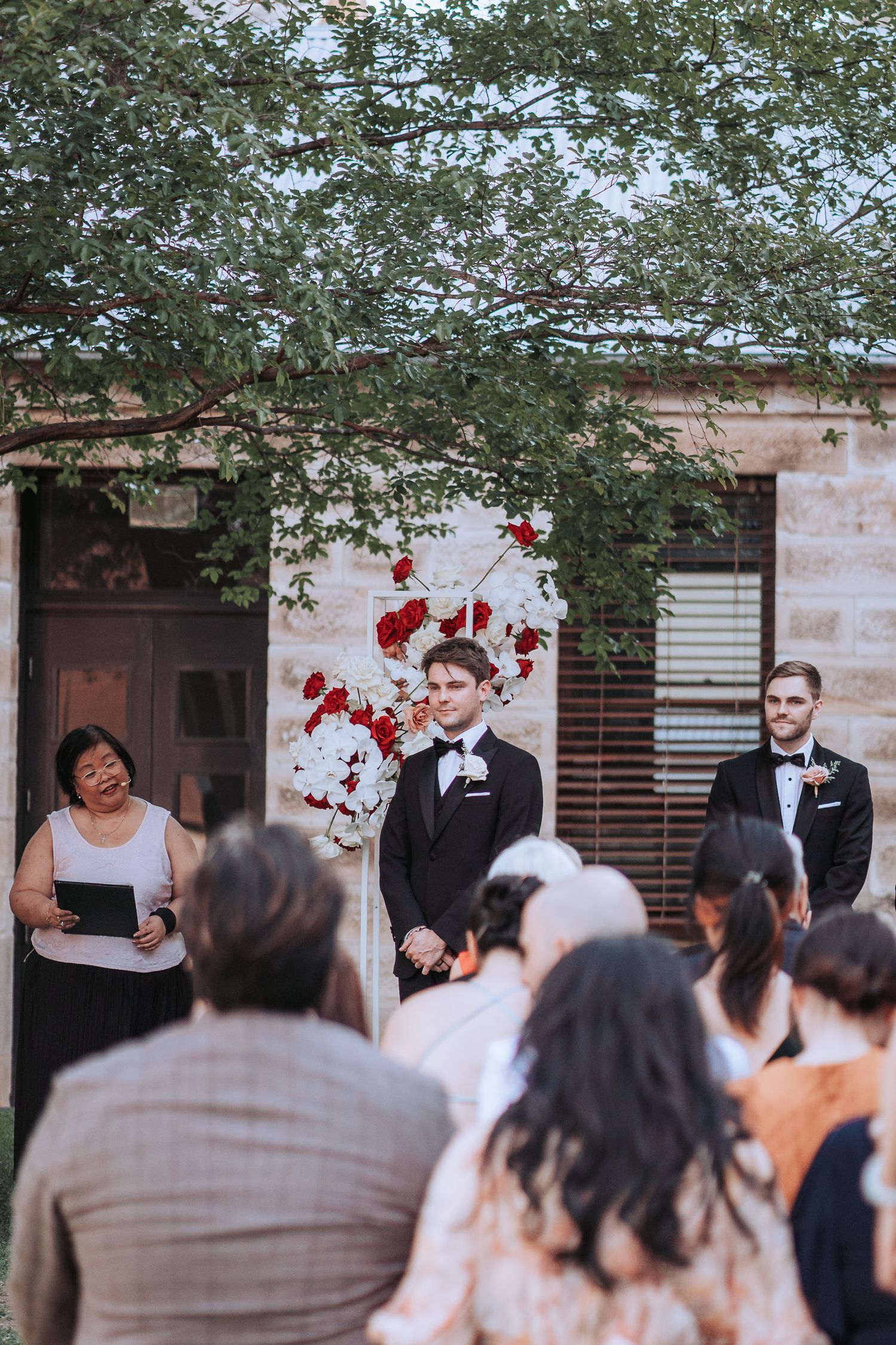 Outdoor wedding ceremony setup with red floral arrangements and guests gathered under trees.