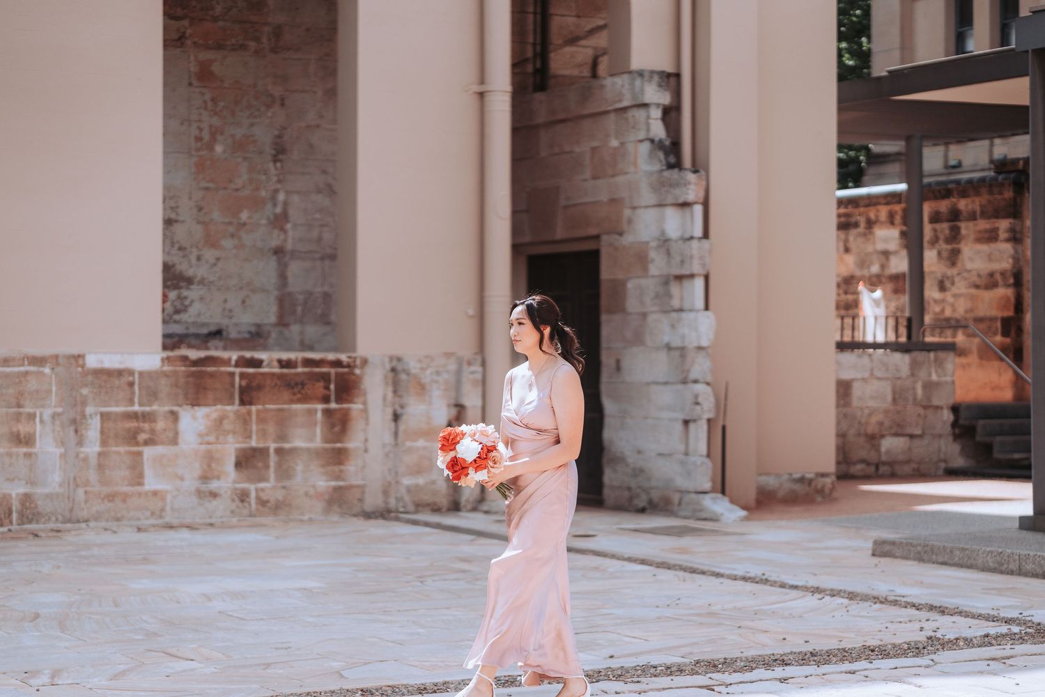 Woman in pink dress holds bouquet while walking along stone building exterior.