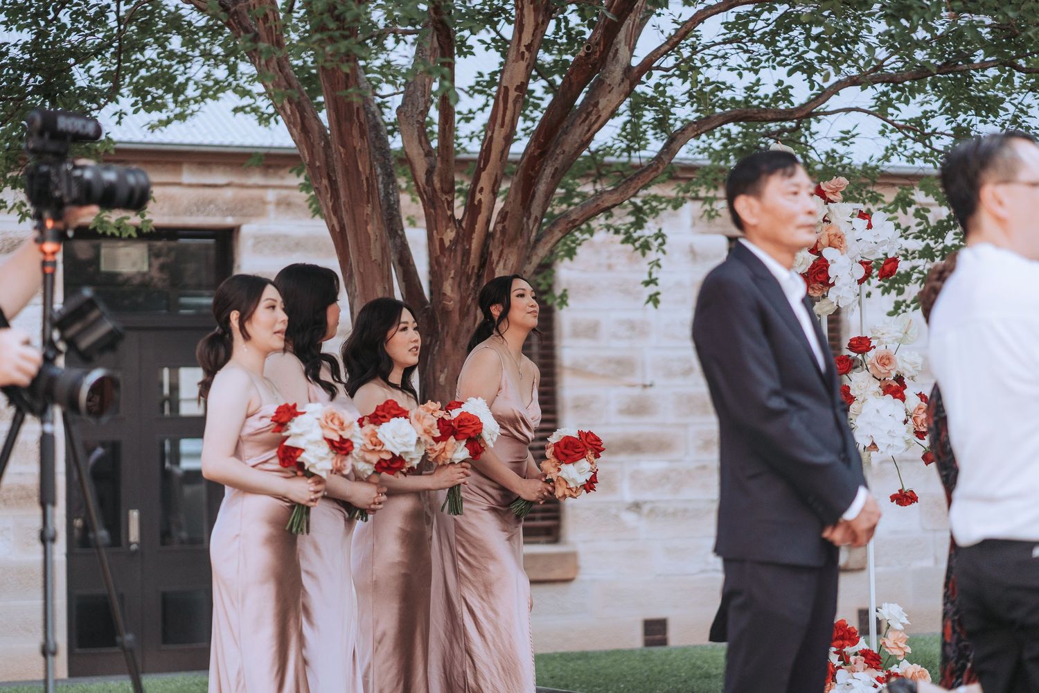 Wedding party members in pink dresses hold red bouquets during outdoor ceremony.