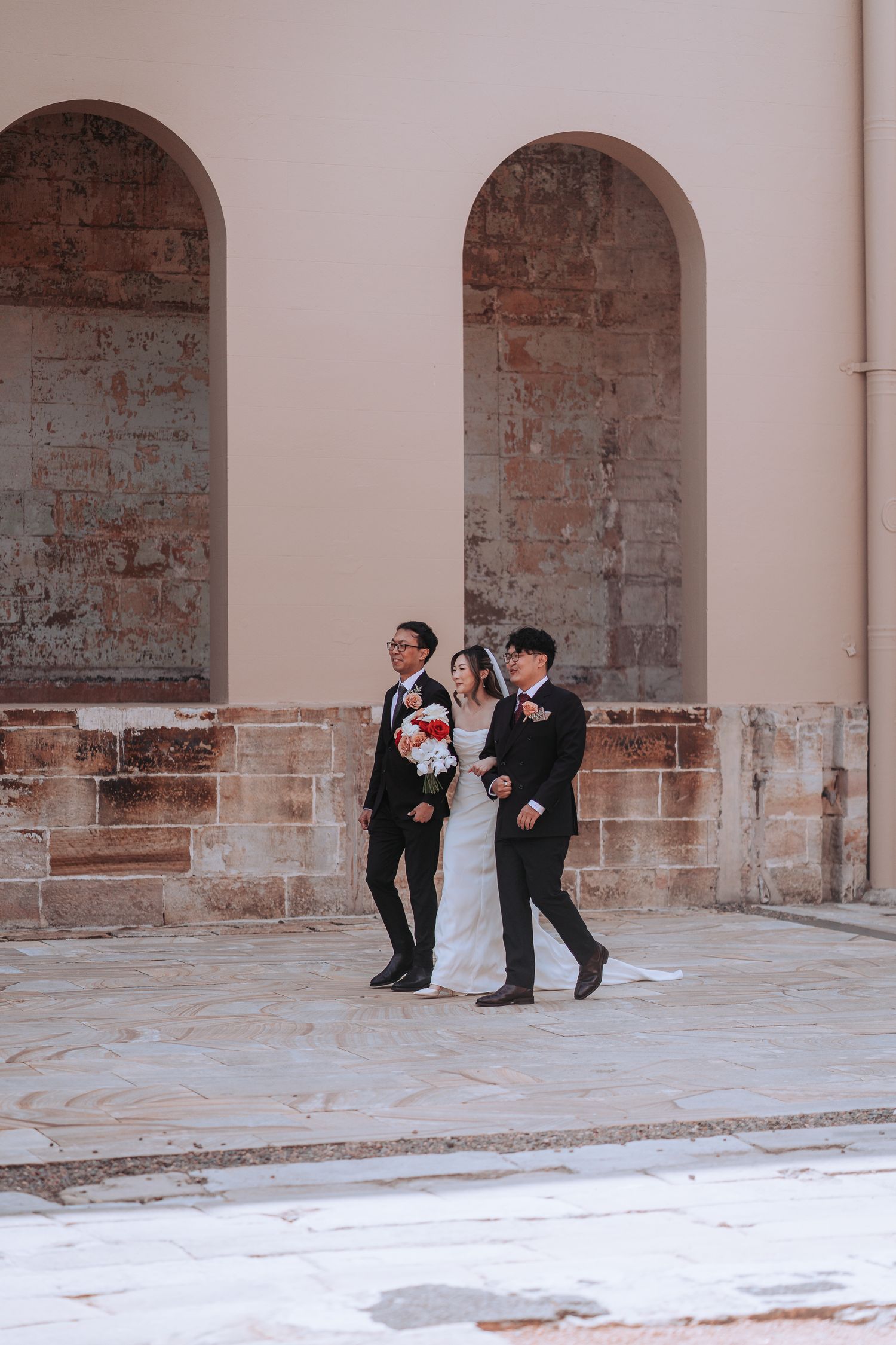 Three people in formal attire walk through stone archways at wedding venue.
