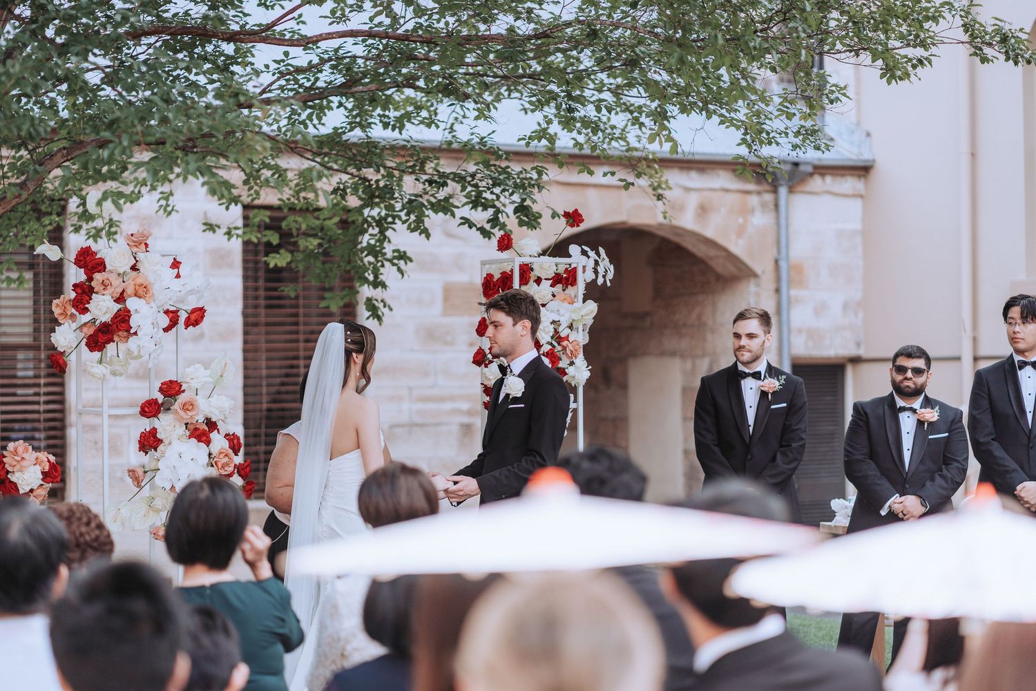 Outdoor wedding ceremony with red floral decor and tree canopy overhead.