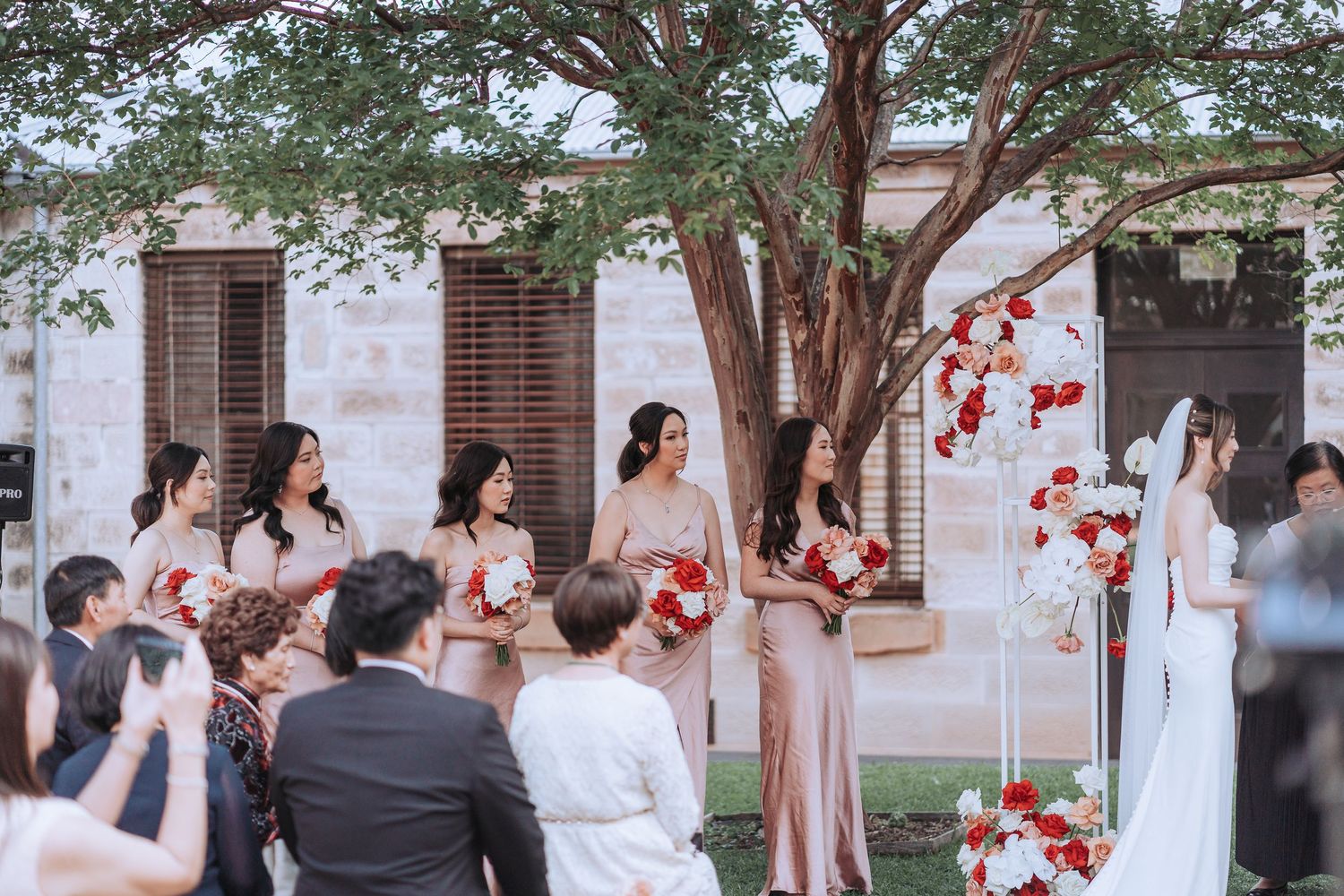Wedding ceremony gathering with bridesmaids in pink dresses and red floral decorations.