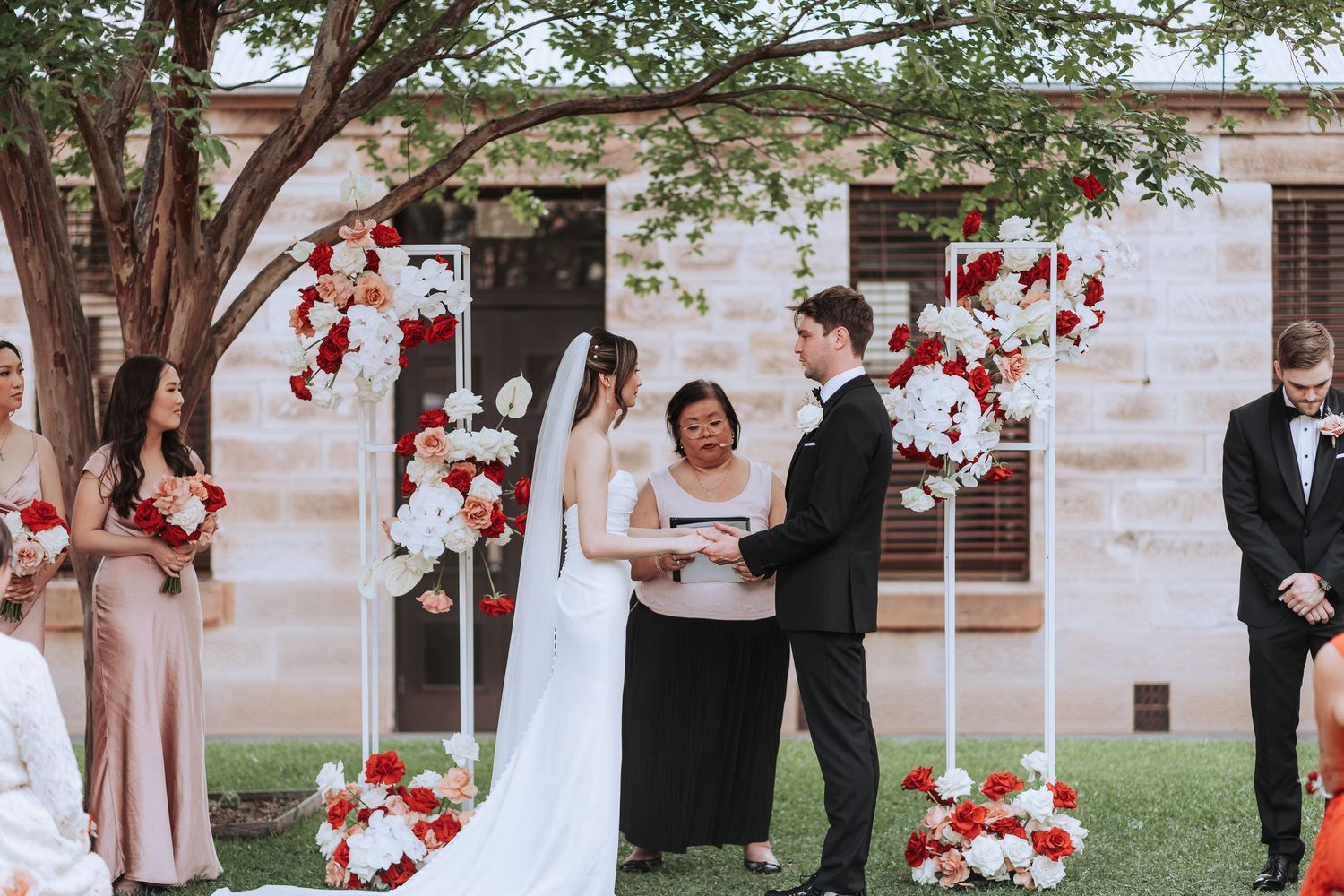 Outdoor wedding ceremony under a tree with red and white floral arrangements and stone building backdrop.