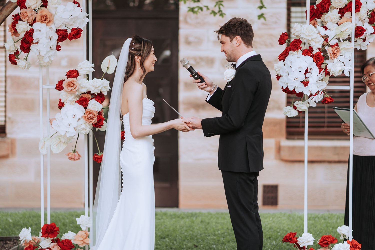 Couple reading vows during outdoor ceremony with red and white floral arch decorations.