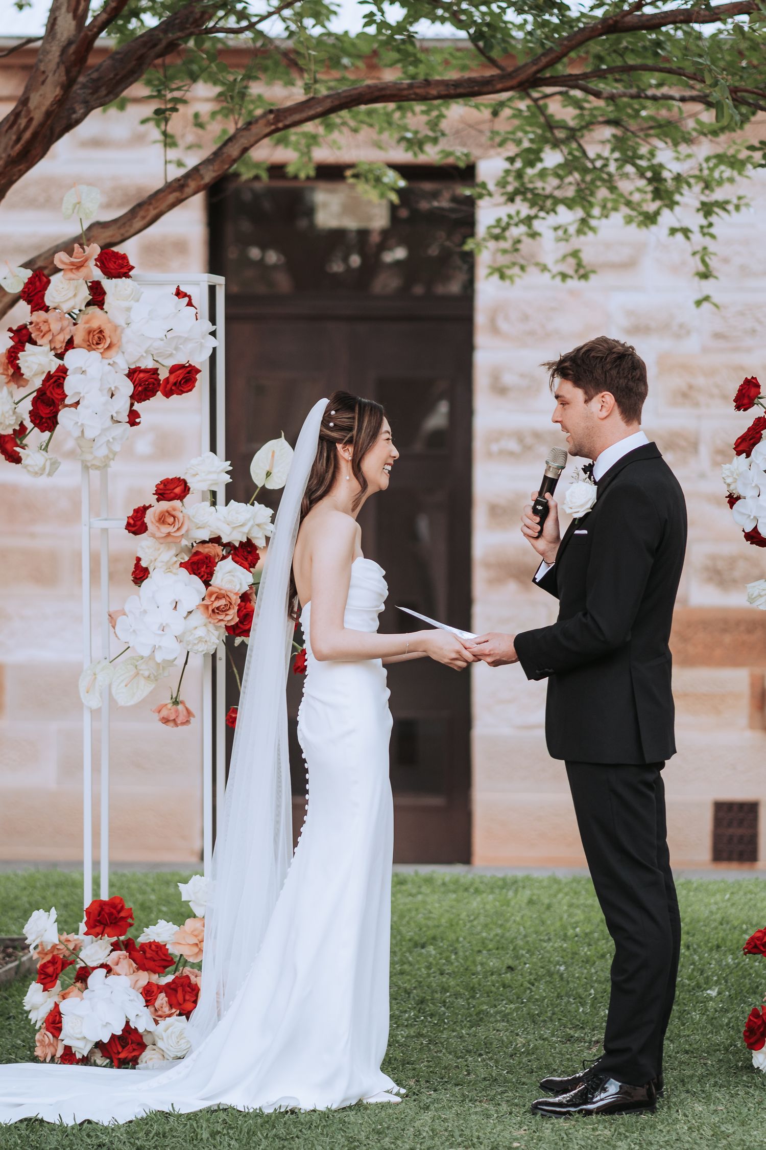 Outdoor wedding ceremony with couple exchanging vows under tree with red and white flower arrangements.