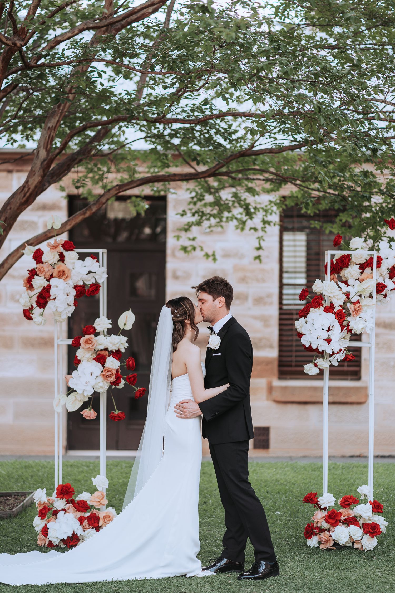 Wedding couple embracing under tree with elegant red and white floral arrangements.
