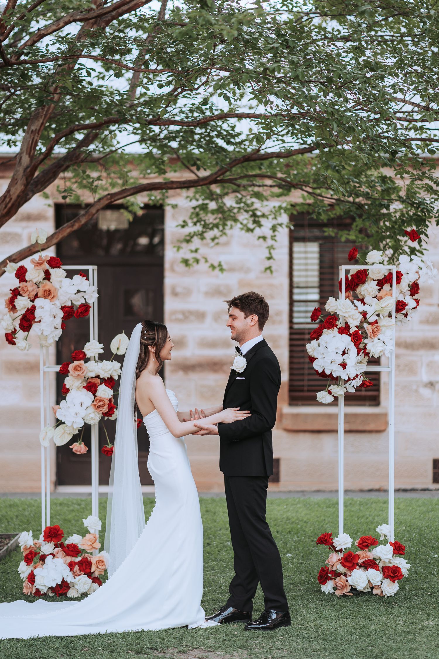 Outdoor wedding ceremony with white and red floral arrangements on stands framing the couple under a tree.