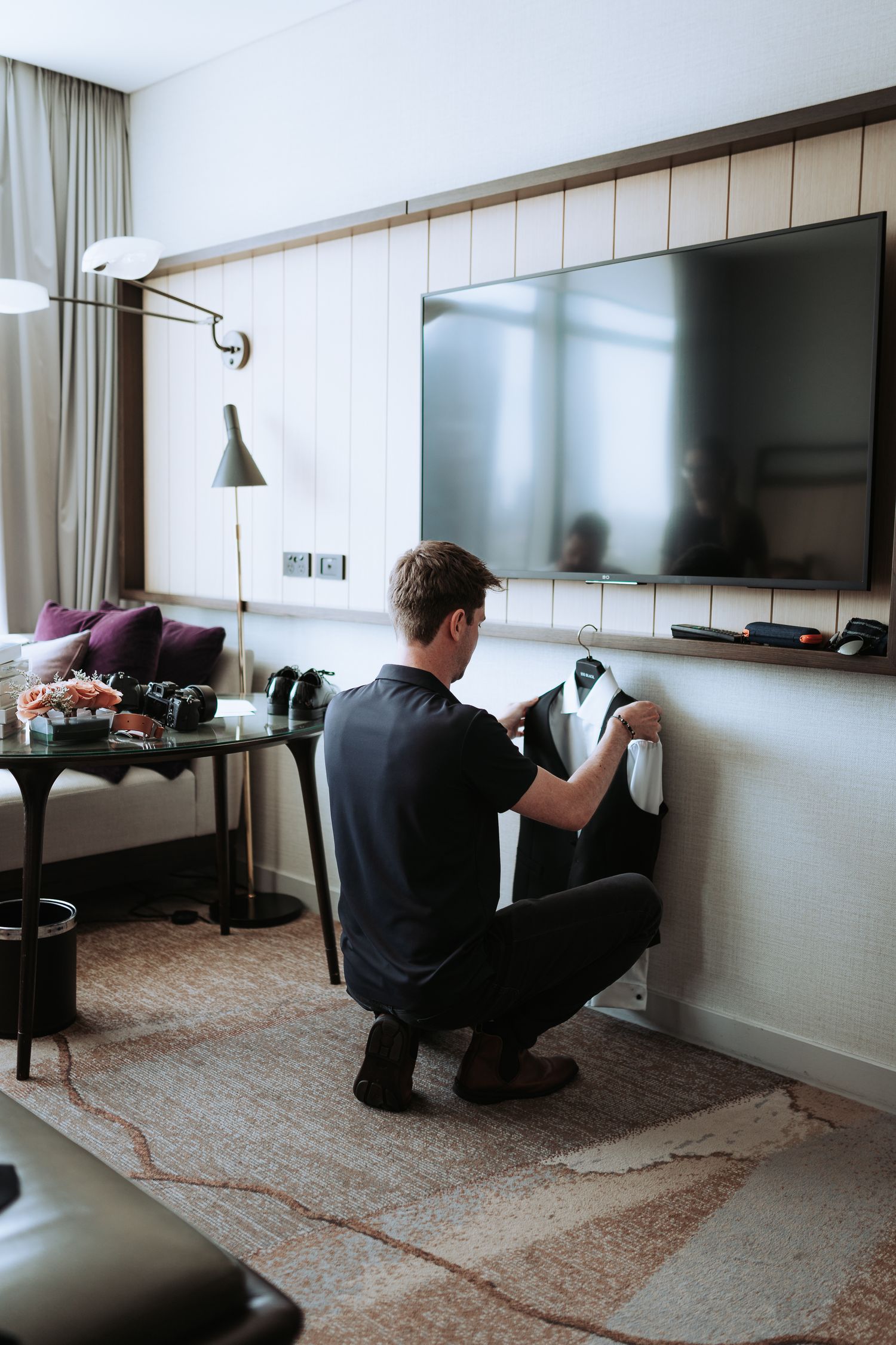 Person in black shirt adjusting formal attire in a modern hotel room with mounted TV.