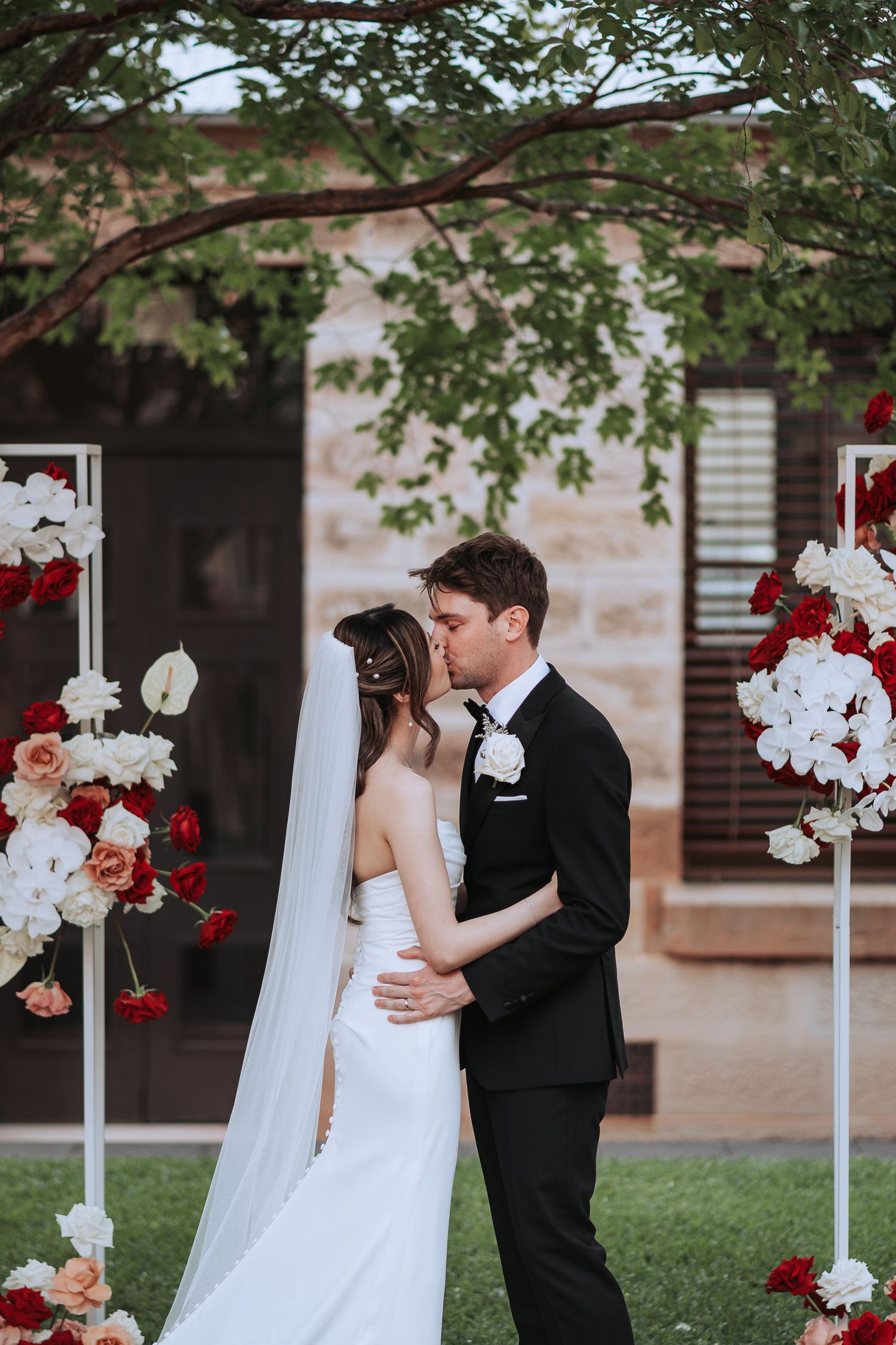 Intimate wedding moment captured under a tree with red and white floral stands framing the scene.