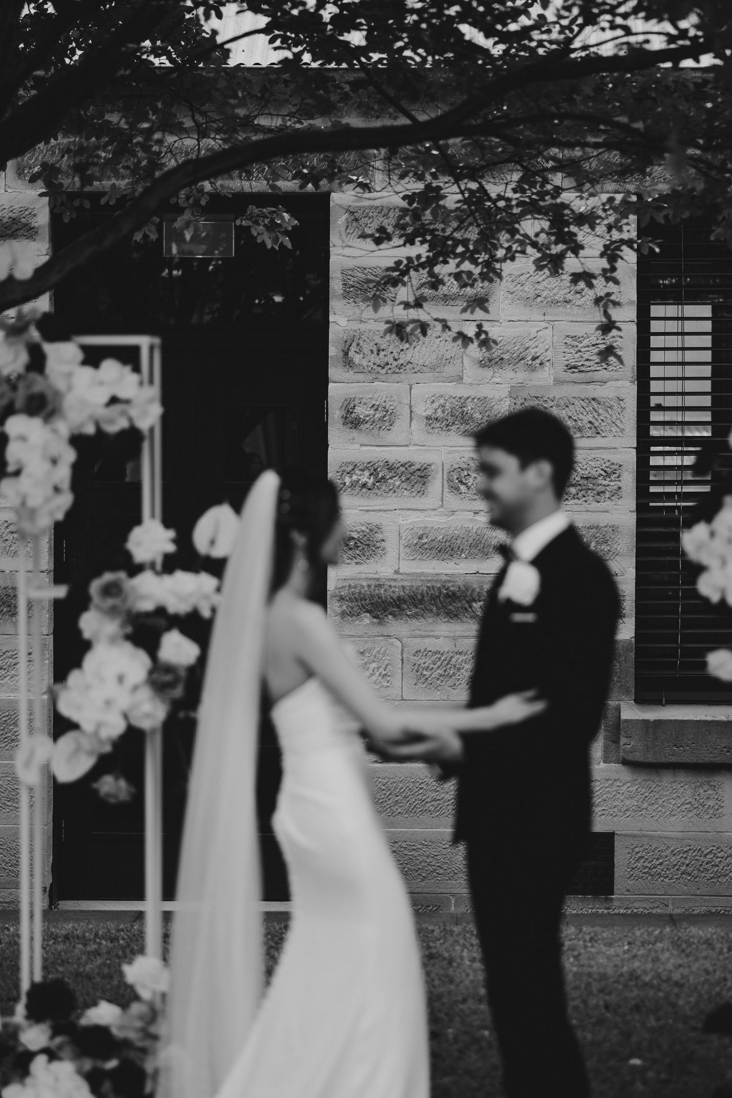 Black and white wedding photo showing couple during ceremony with floral decorations and stone building backdrop.