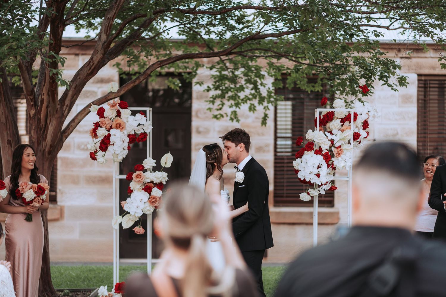 Wedding ceremony under a tree with red and white floral arrangements as guests look on.